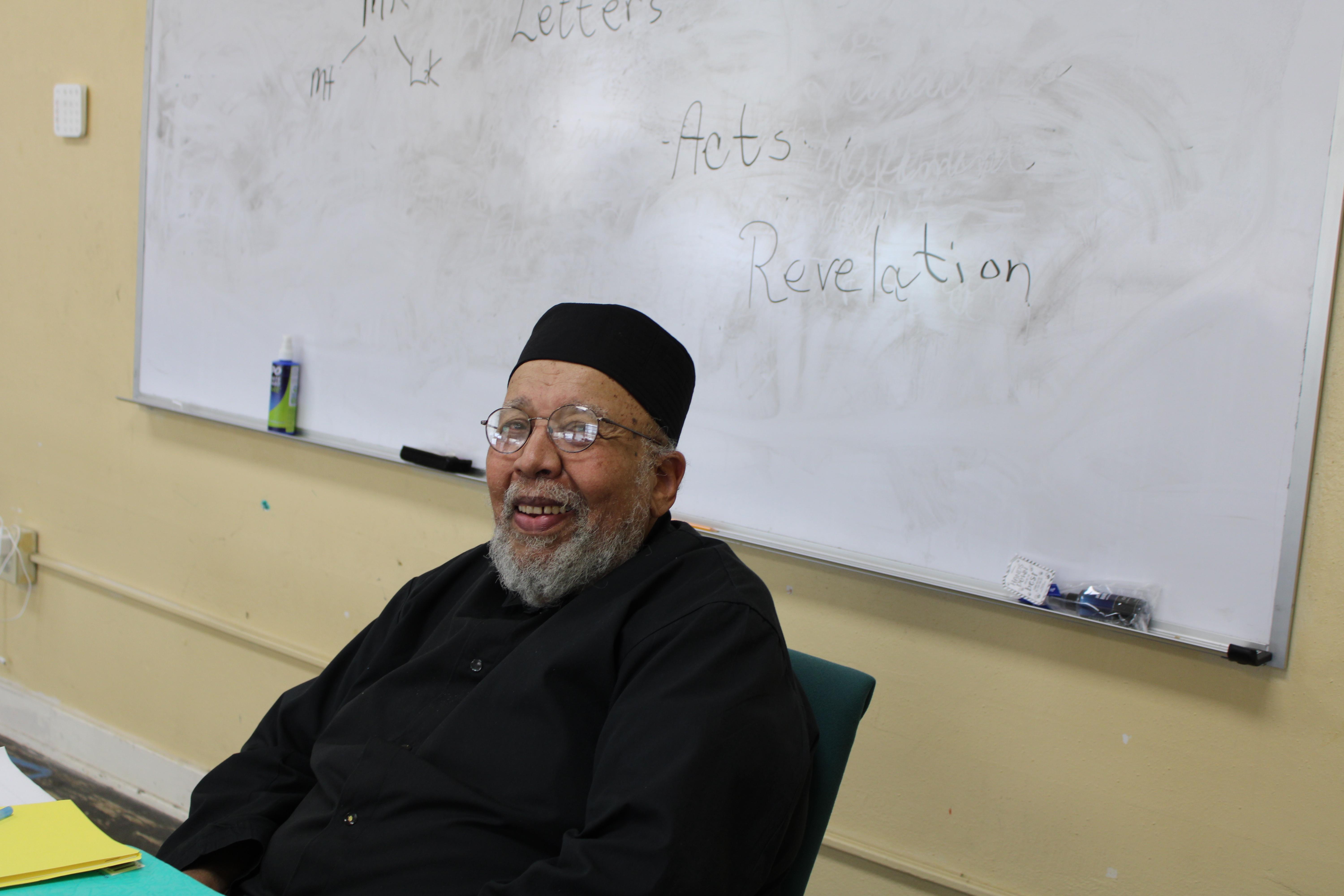 An elder priest dressed in black religious attire sits at a desk.