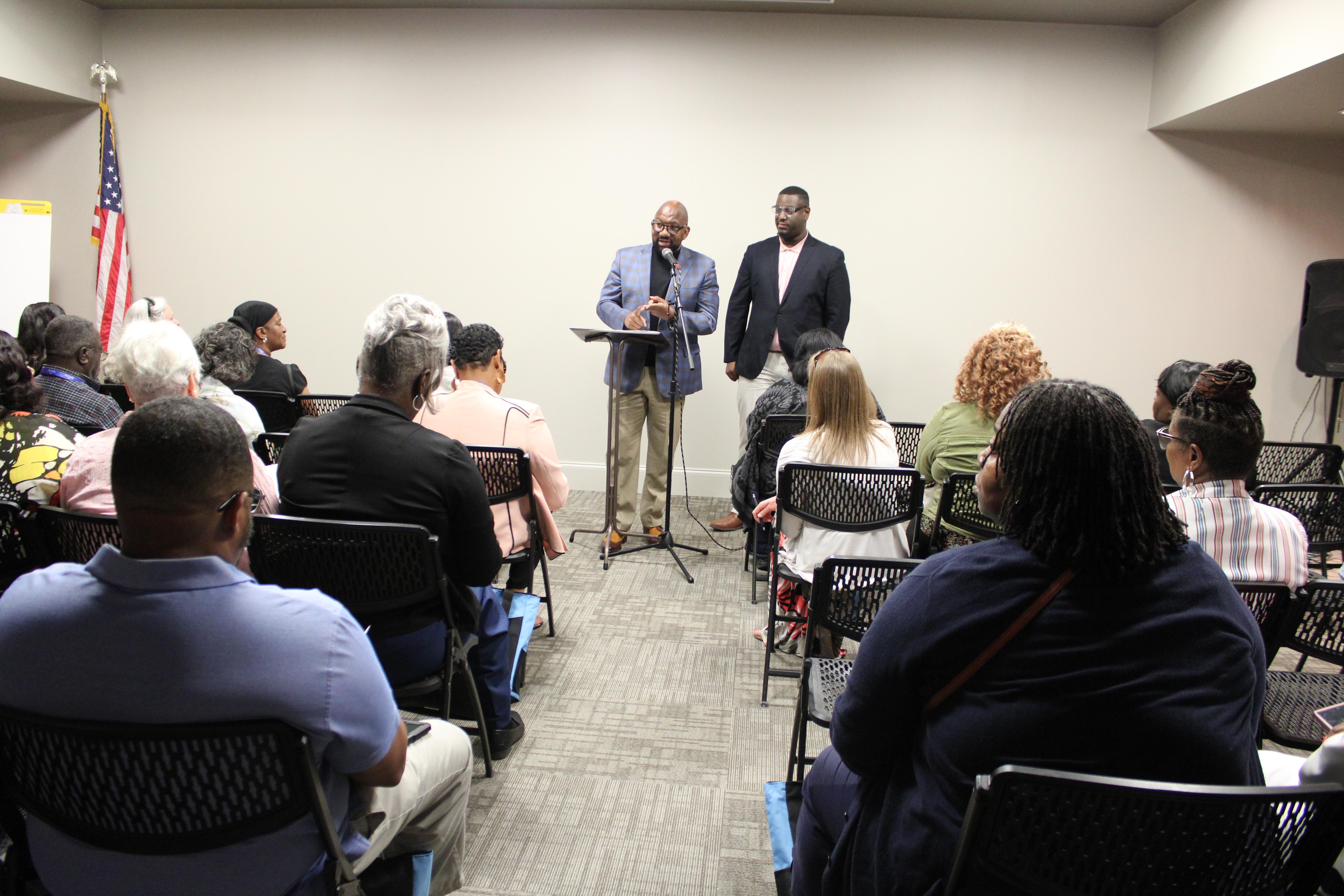 A seated group of people listen as two men speak from a podium.