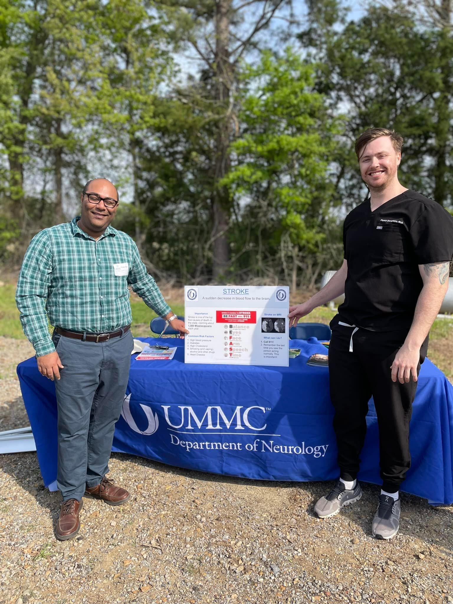 Two men stand before a table draped in a blue sign reading 'UMMC: Department of Neurology.'