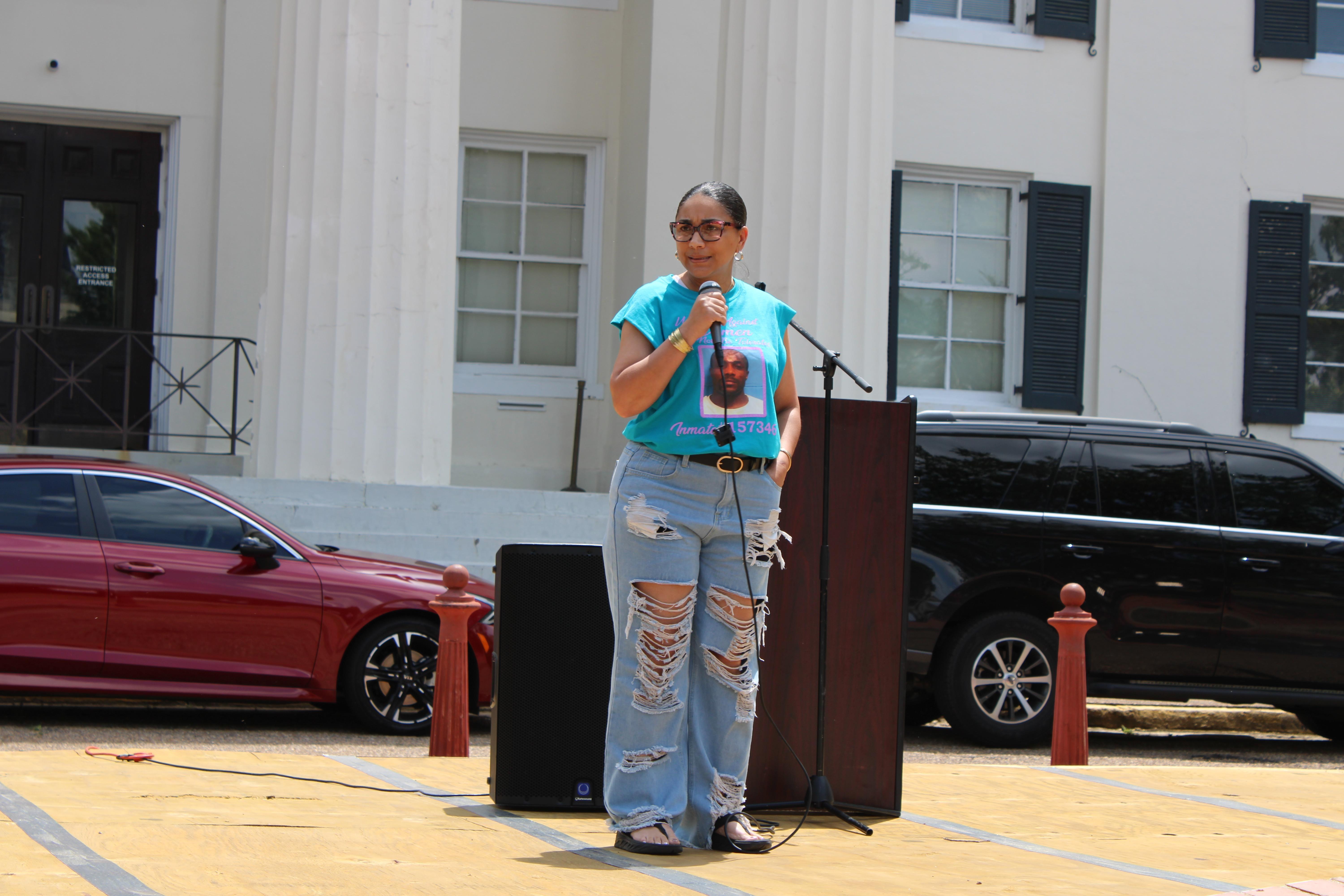 A woman stands at a podium, wearing ripped jeans and a t-shirt with an inmate's mugshot displayed.