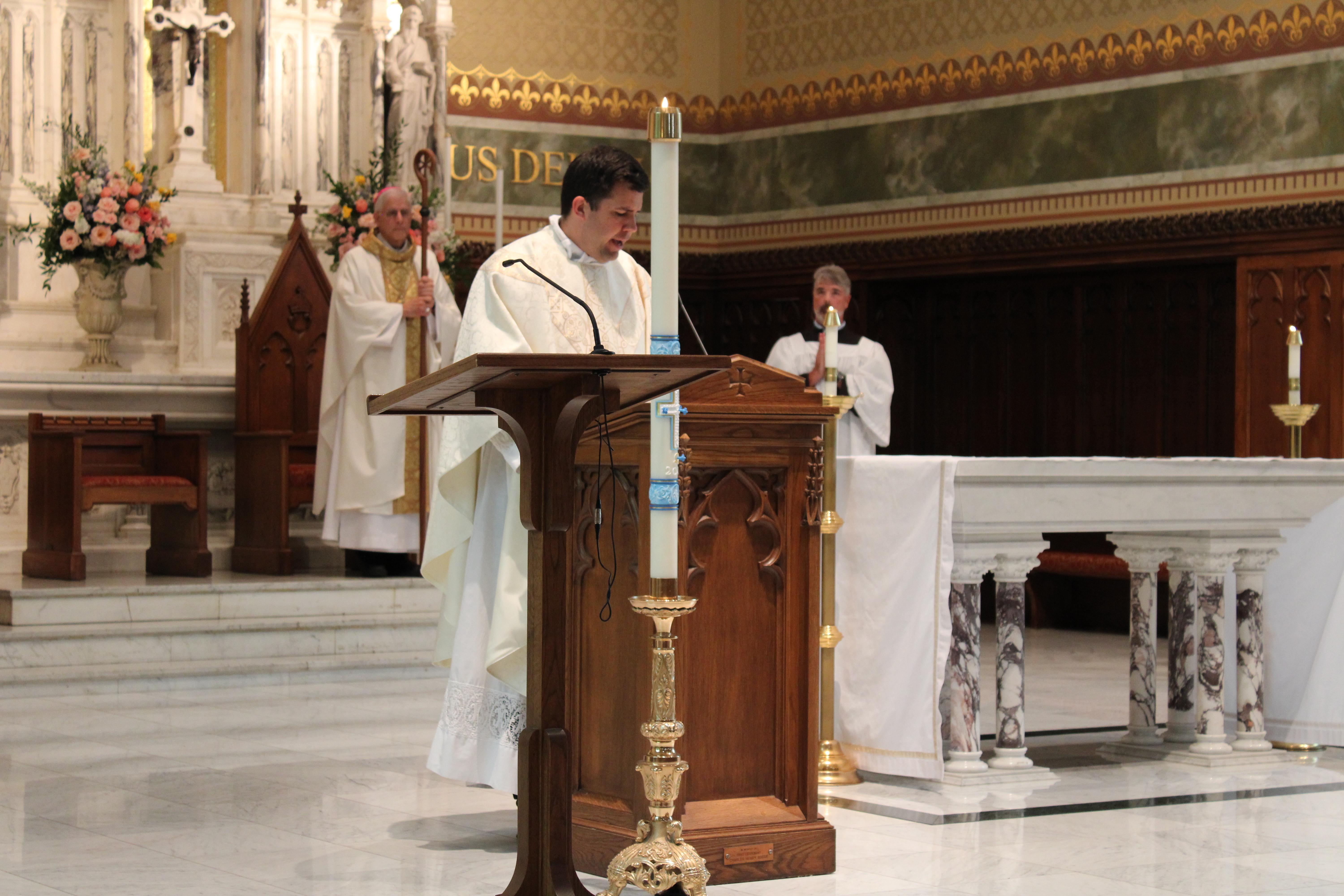 A priest prays at a podium, surrounded by tall candles, during a mass in a cathedral.