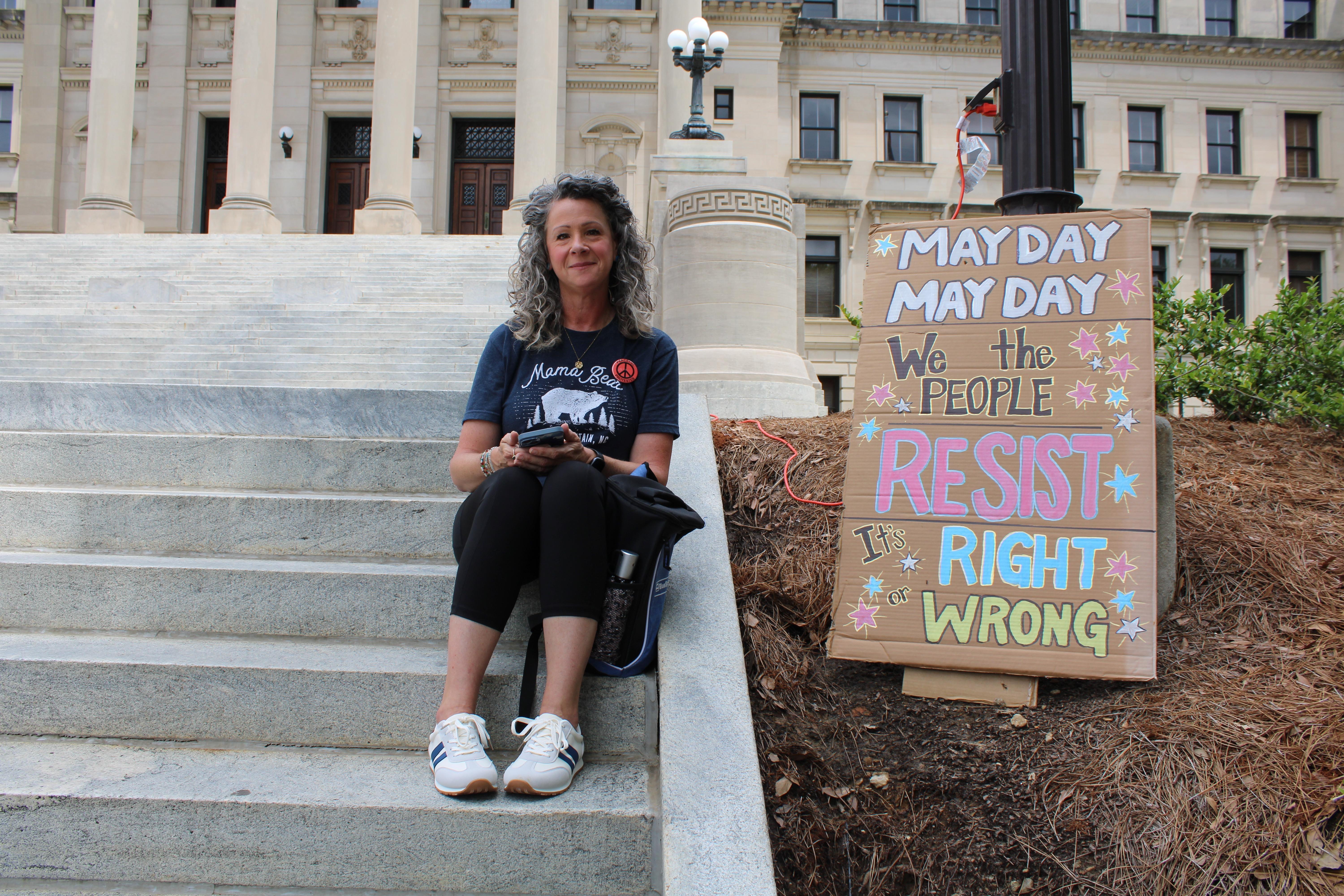 A woman sits next to a sign she created that reads "May Day, we the people resist, it's right or wrong."