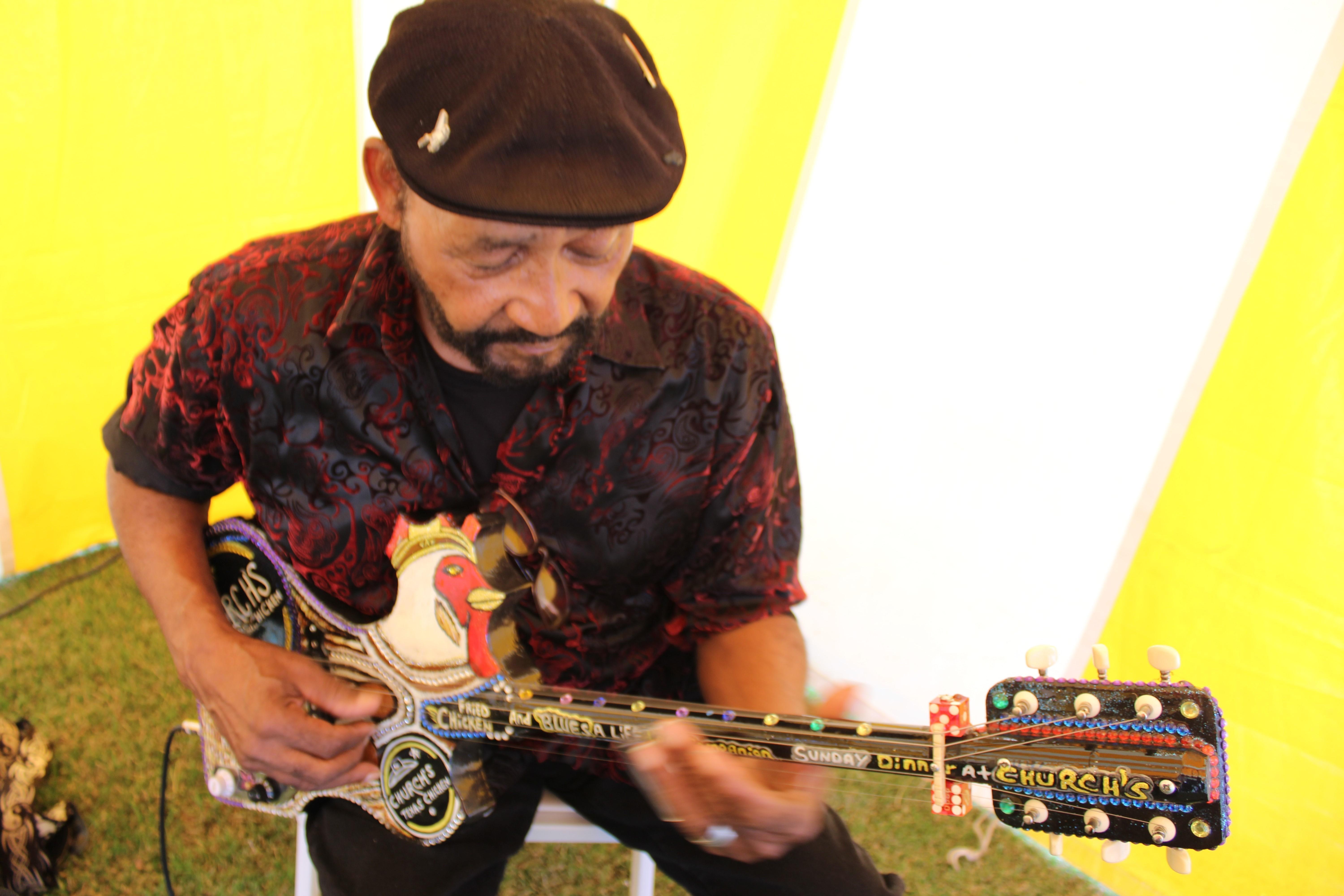A man wearing a black cap and a red, crushed velvet shirt plays a diddly bow made to look like a bejeweled chicken. 