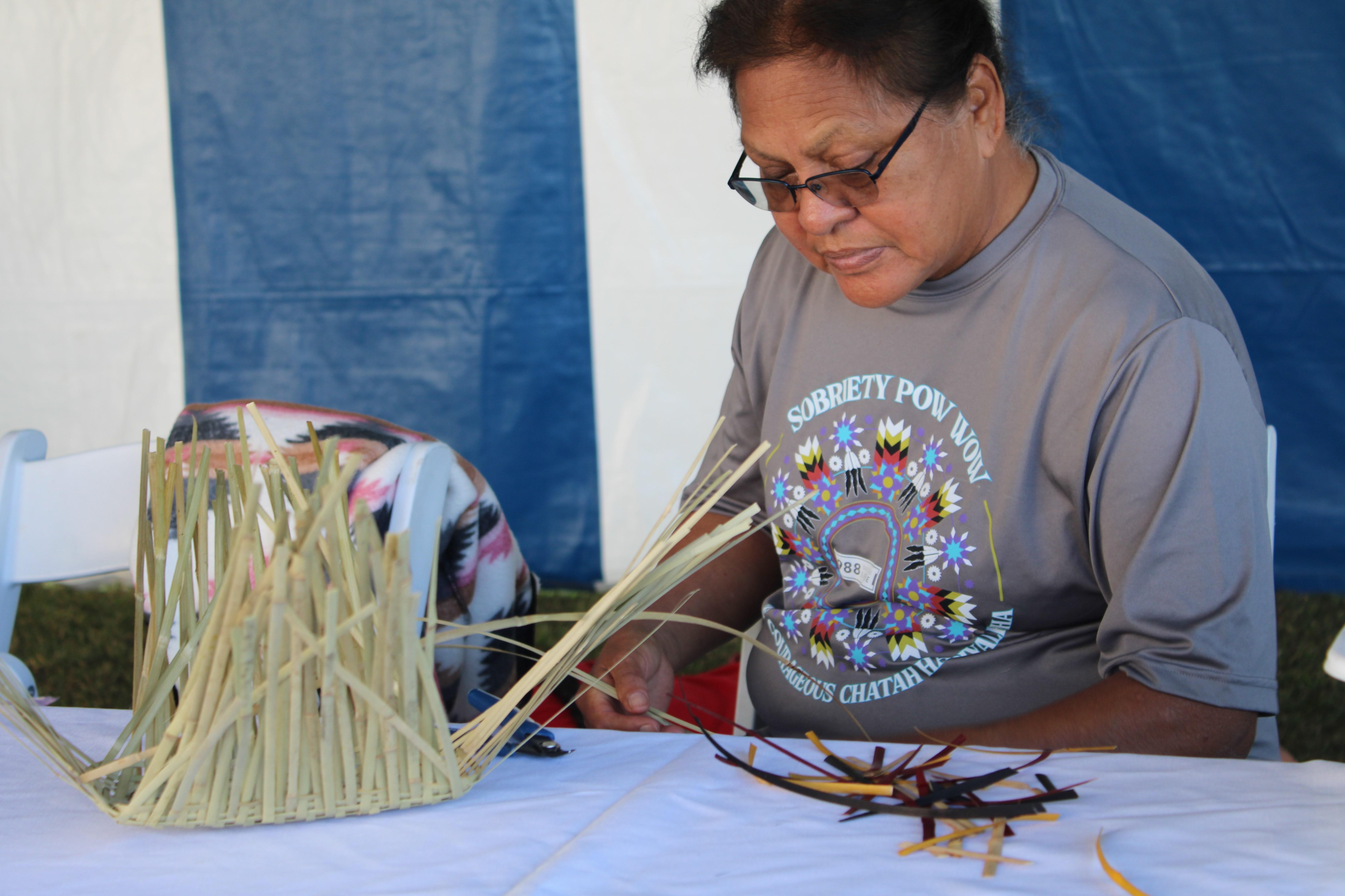 An expert basket weaver sits behind a table that showcases the progress of one tan colored basket and pieces of dyed river cane.
