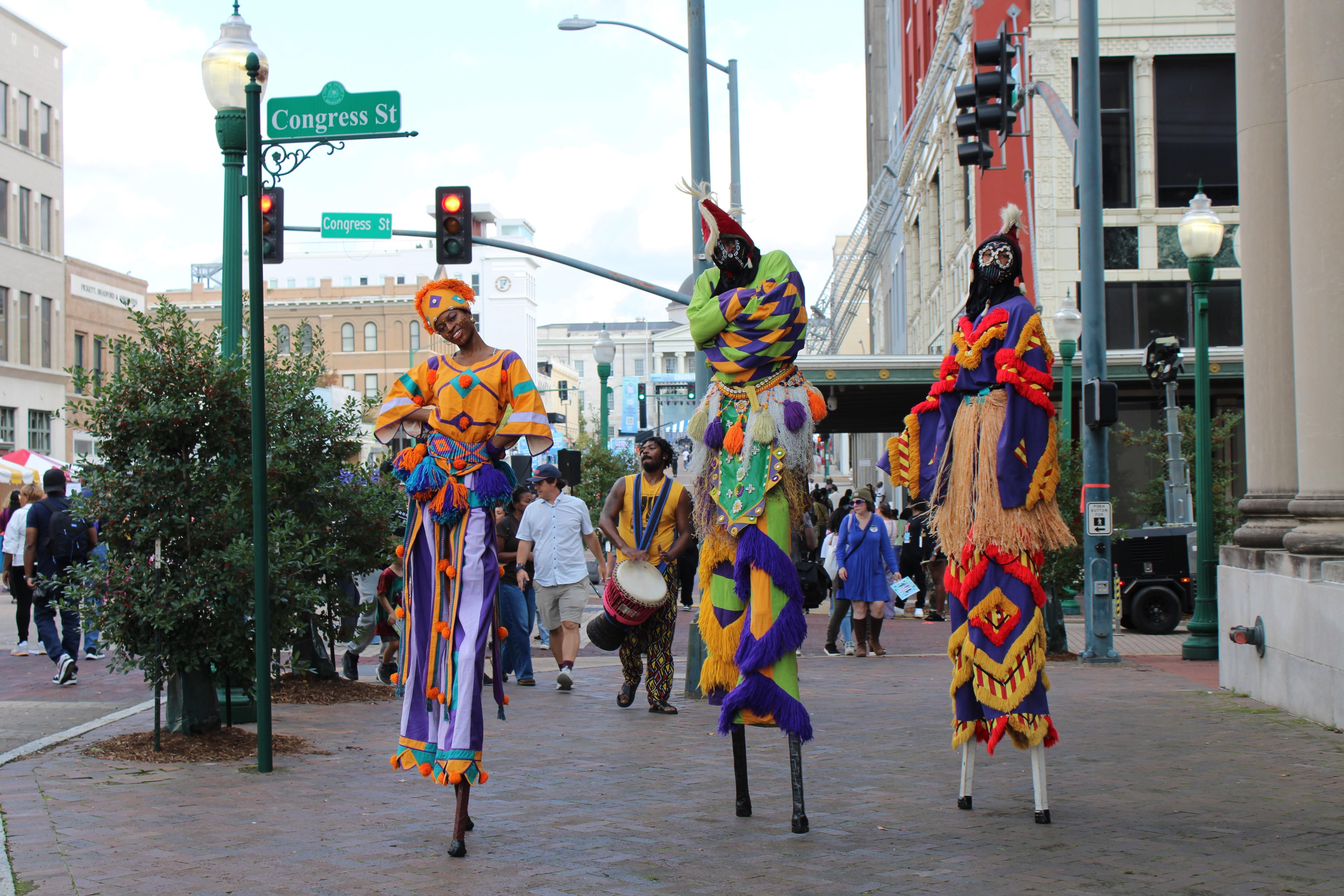 Three stilt dancers dressed in bright, bold African inspired attire, two wearing black masks, pose in the middle of a sidewalk with a crowd seen behind them.