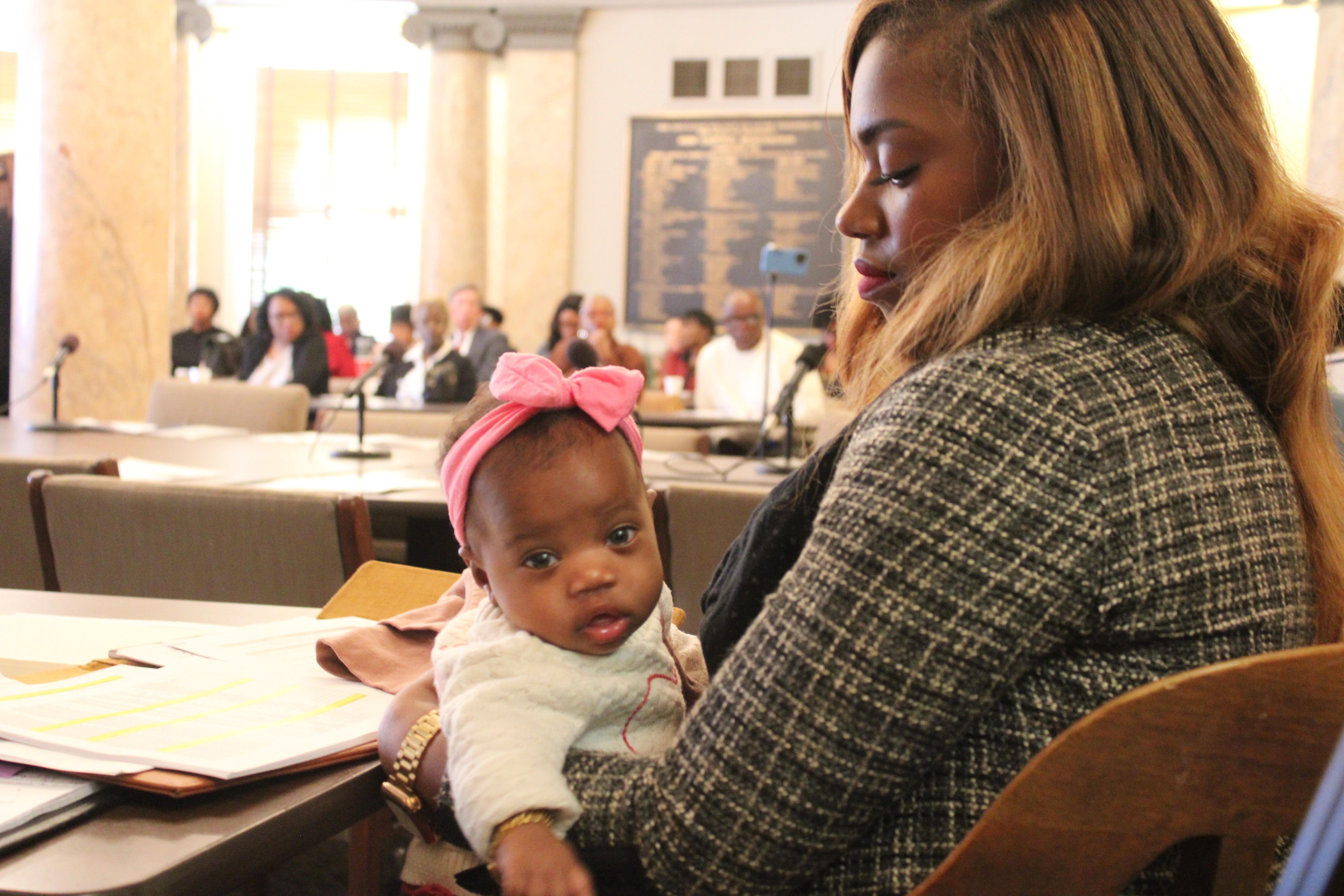 A baby wearing a pink bow looks directly at the camera while being held by her mother.