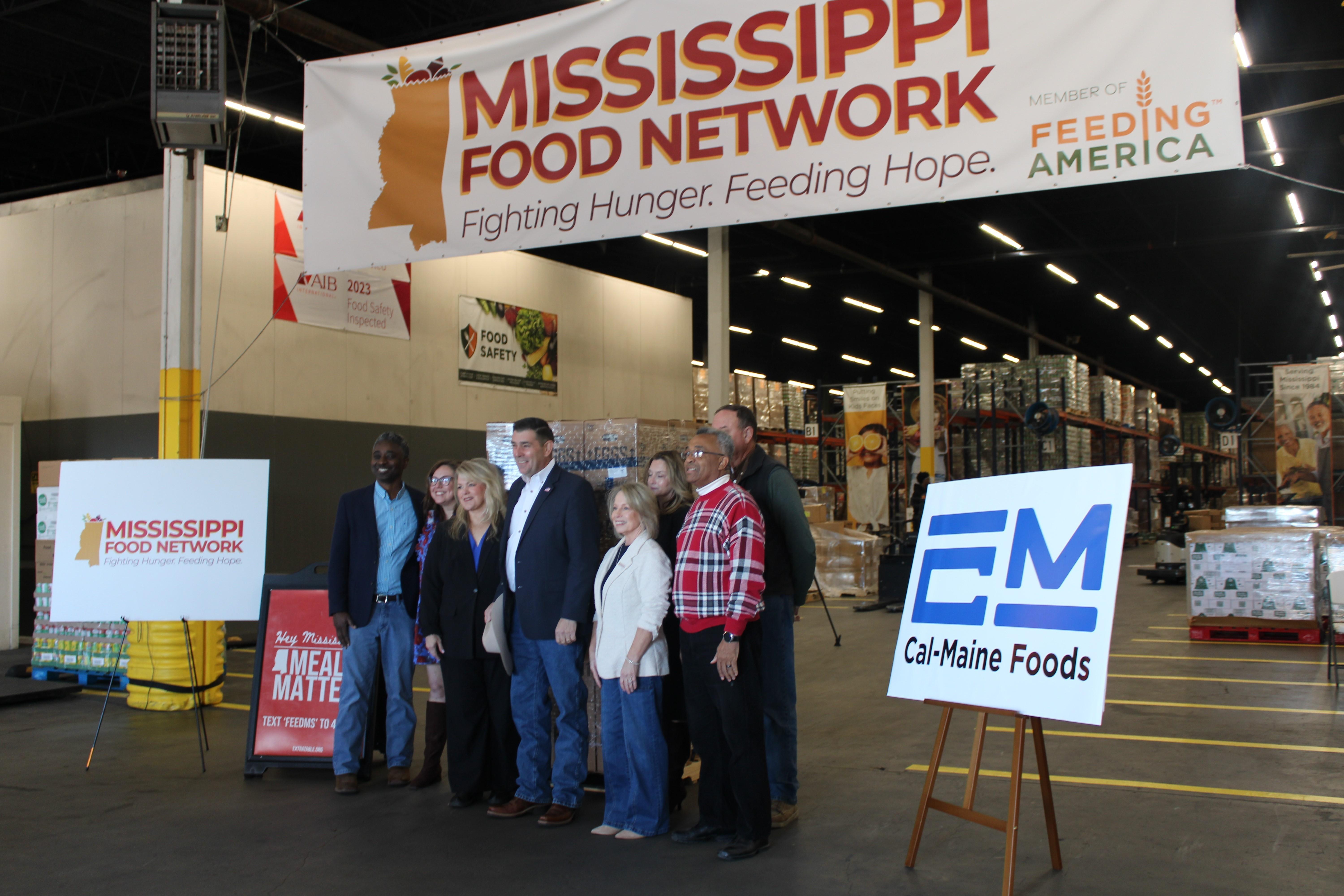 Eight people pose in front a food bank distribution warehouse, smiling.