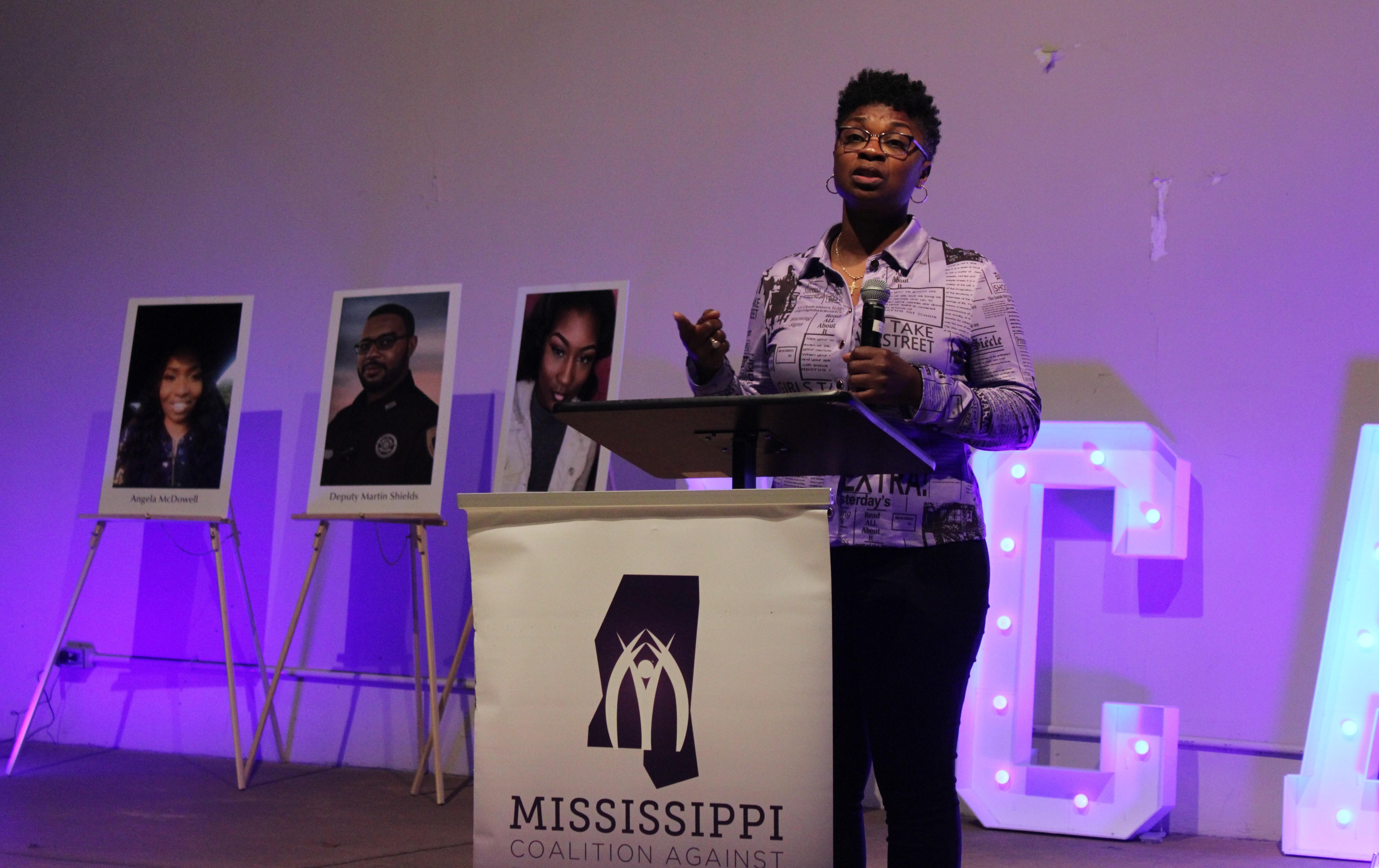 A mother holds a mic while standing in front of a photo of her deceased daughter.