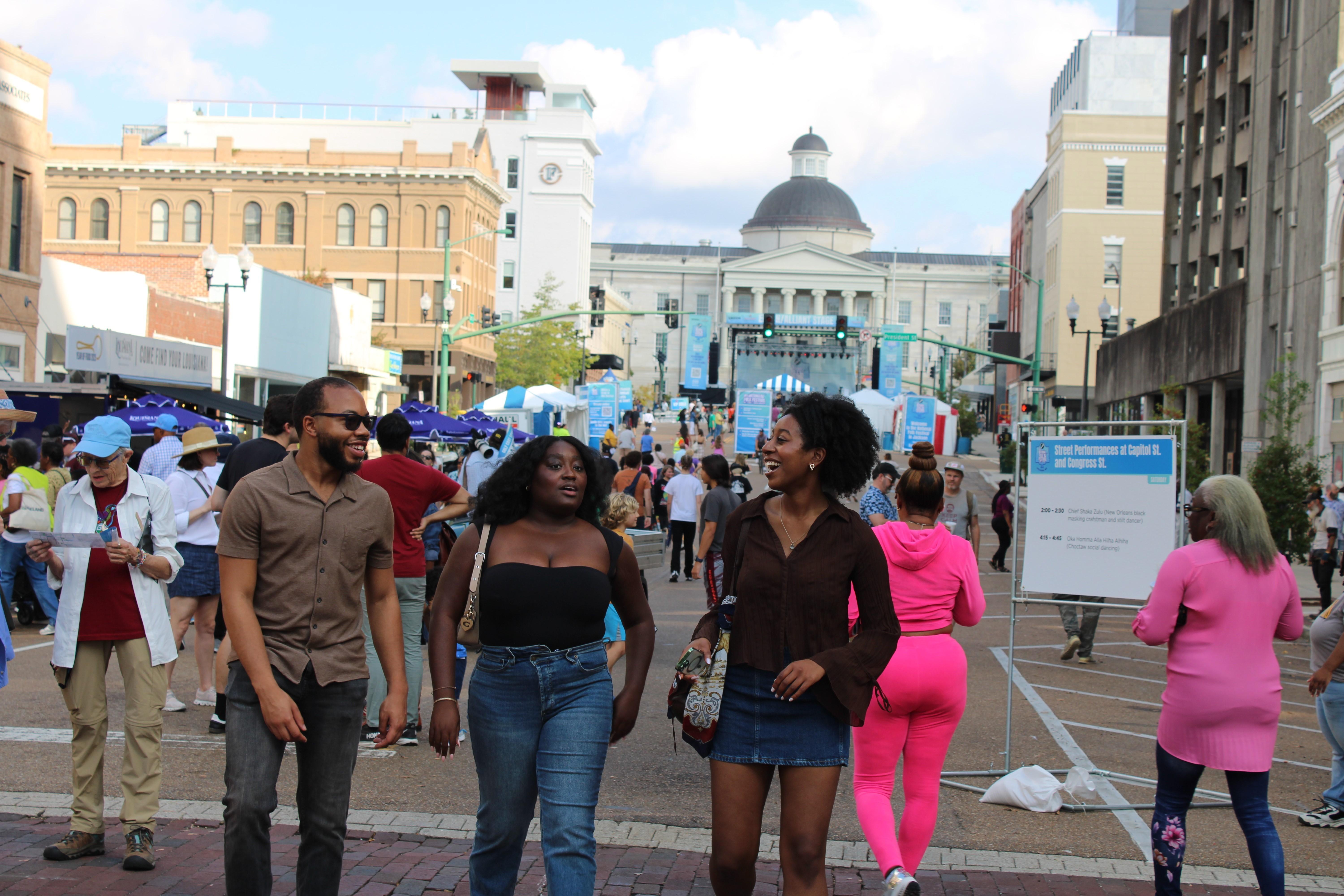 Smiling visitors walk through a busy, festival filled downtown scene.