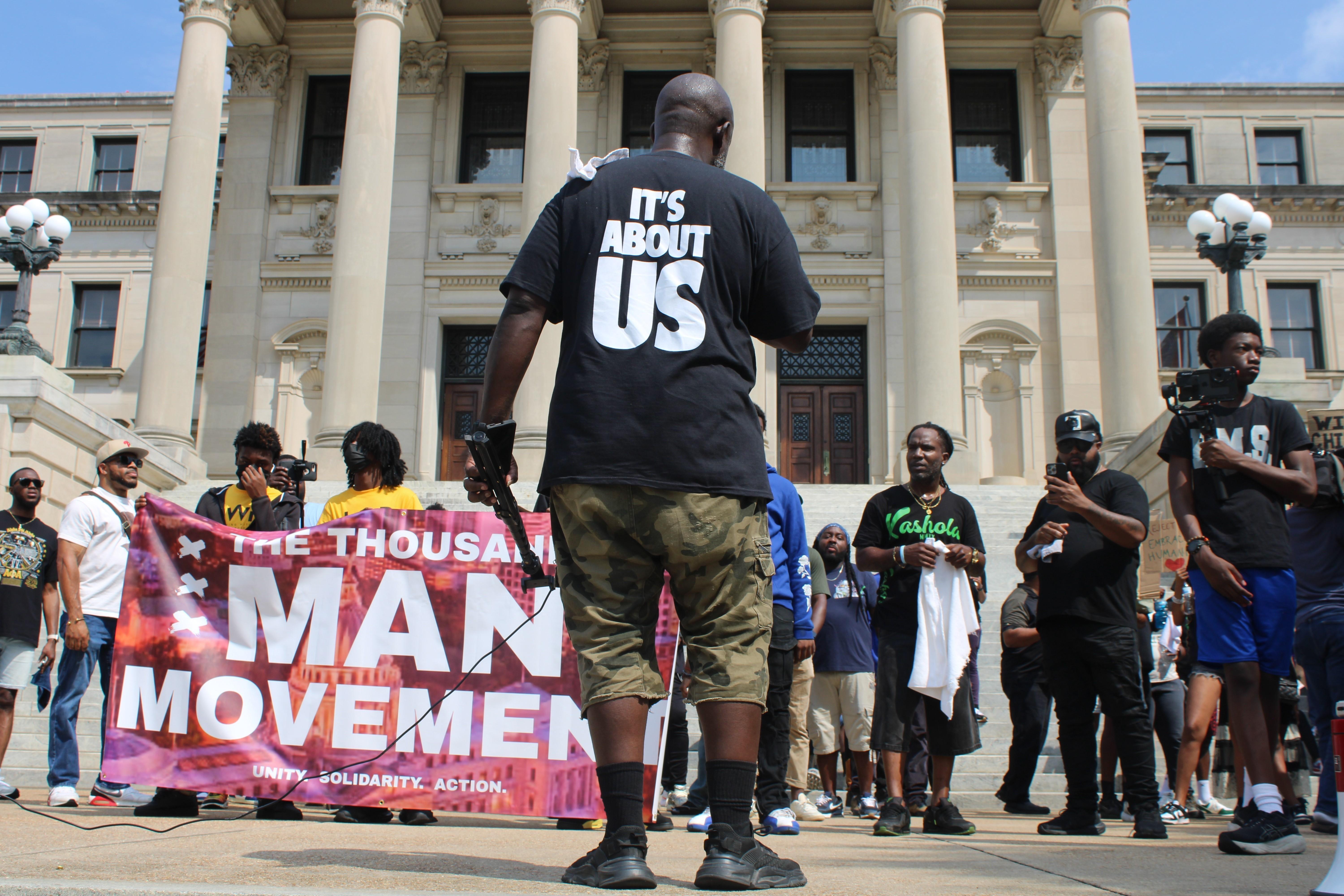 A rally of Black men and younger adolescents hold signs and stand before the Mississippi state capitol building.