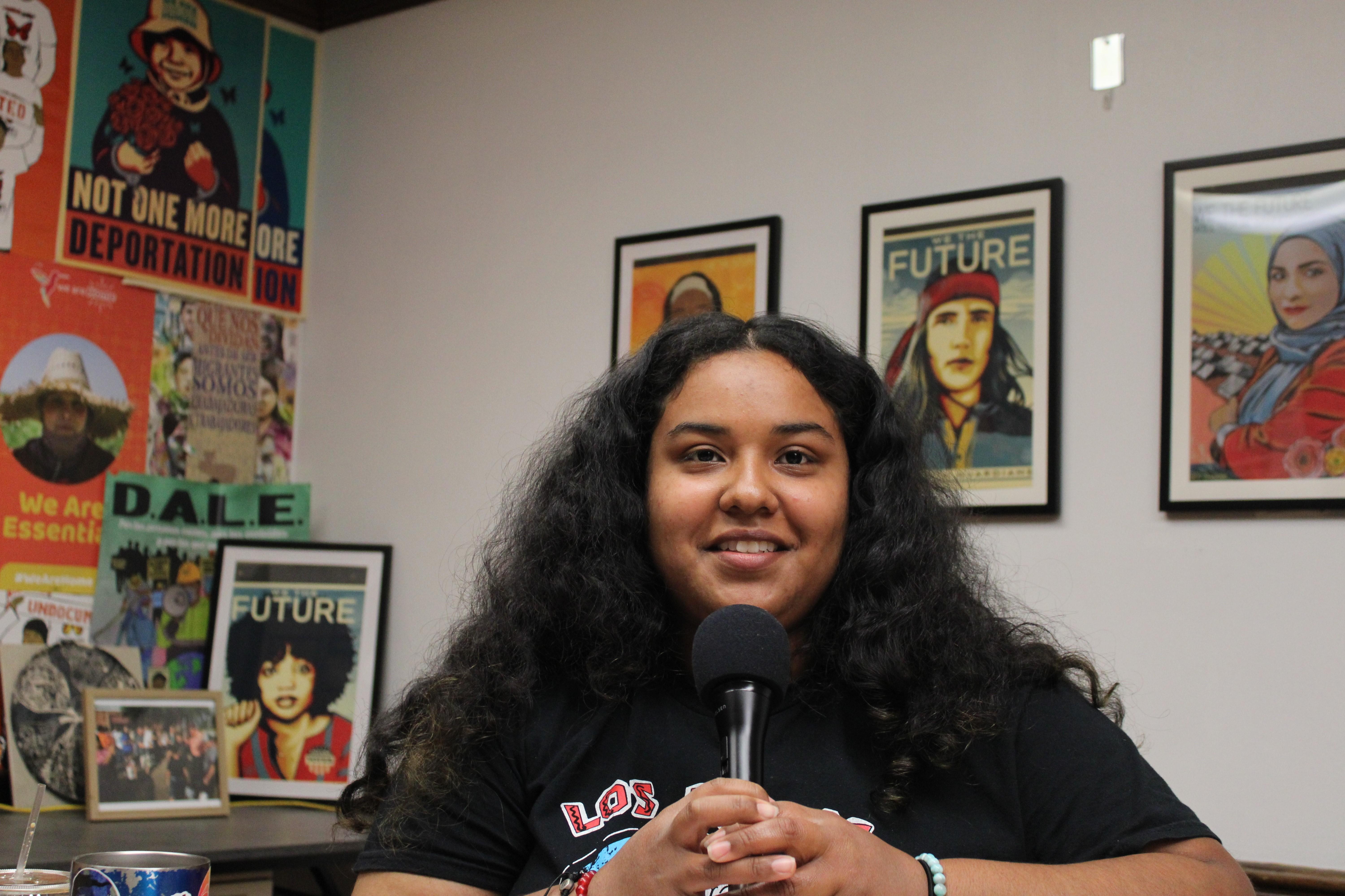 A young woman with full hair speaks into a microphone. Colorful posters are seen in the background.