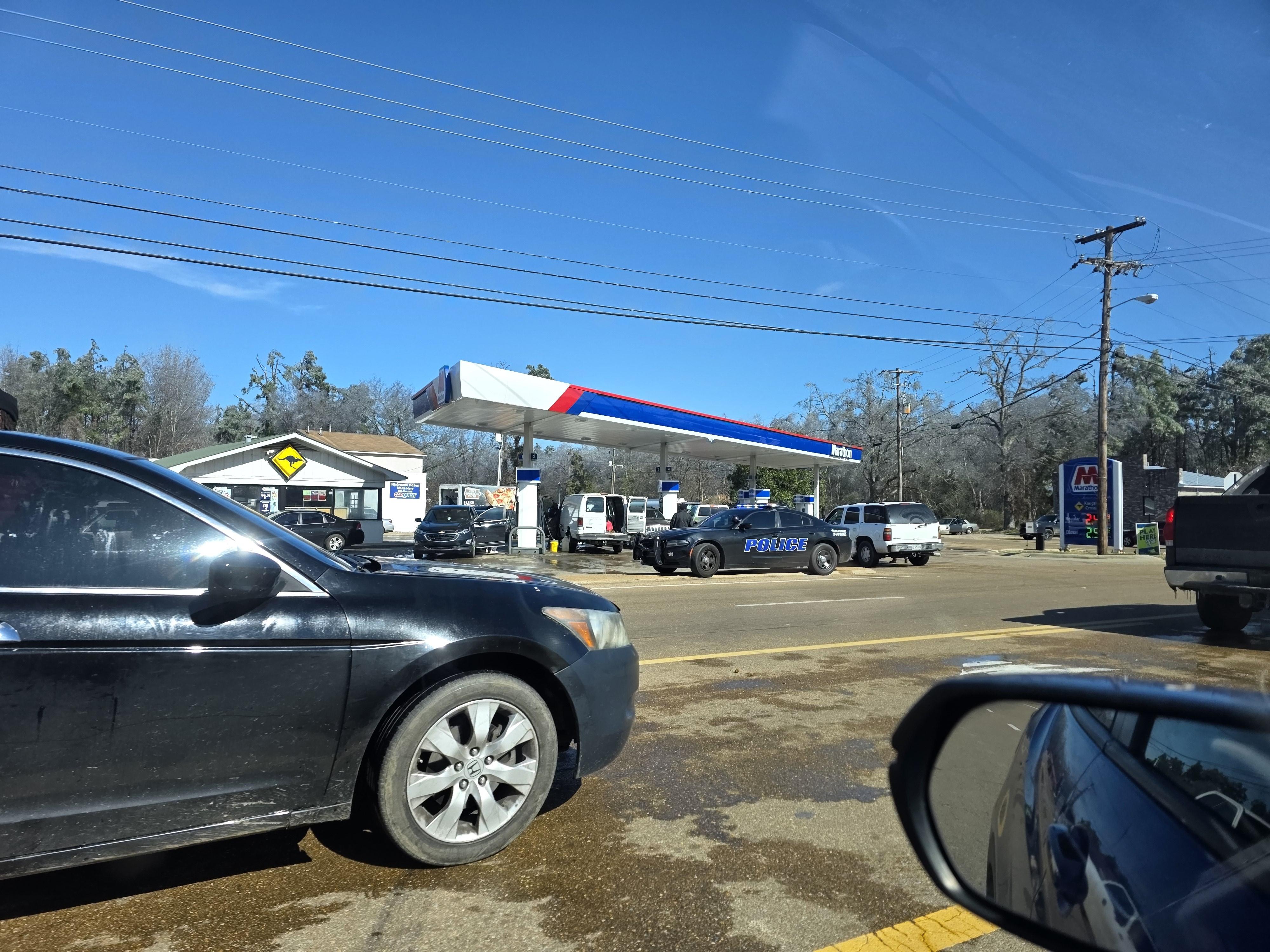 Cars are parked in the median lane of a busy roadway waiting to turn into the gas station. A police cruiser is stationed to help direct traffic.