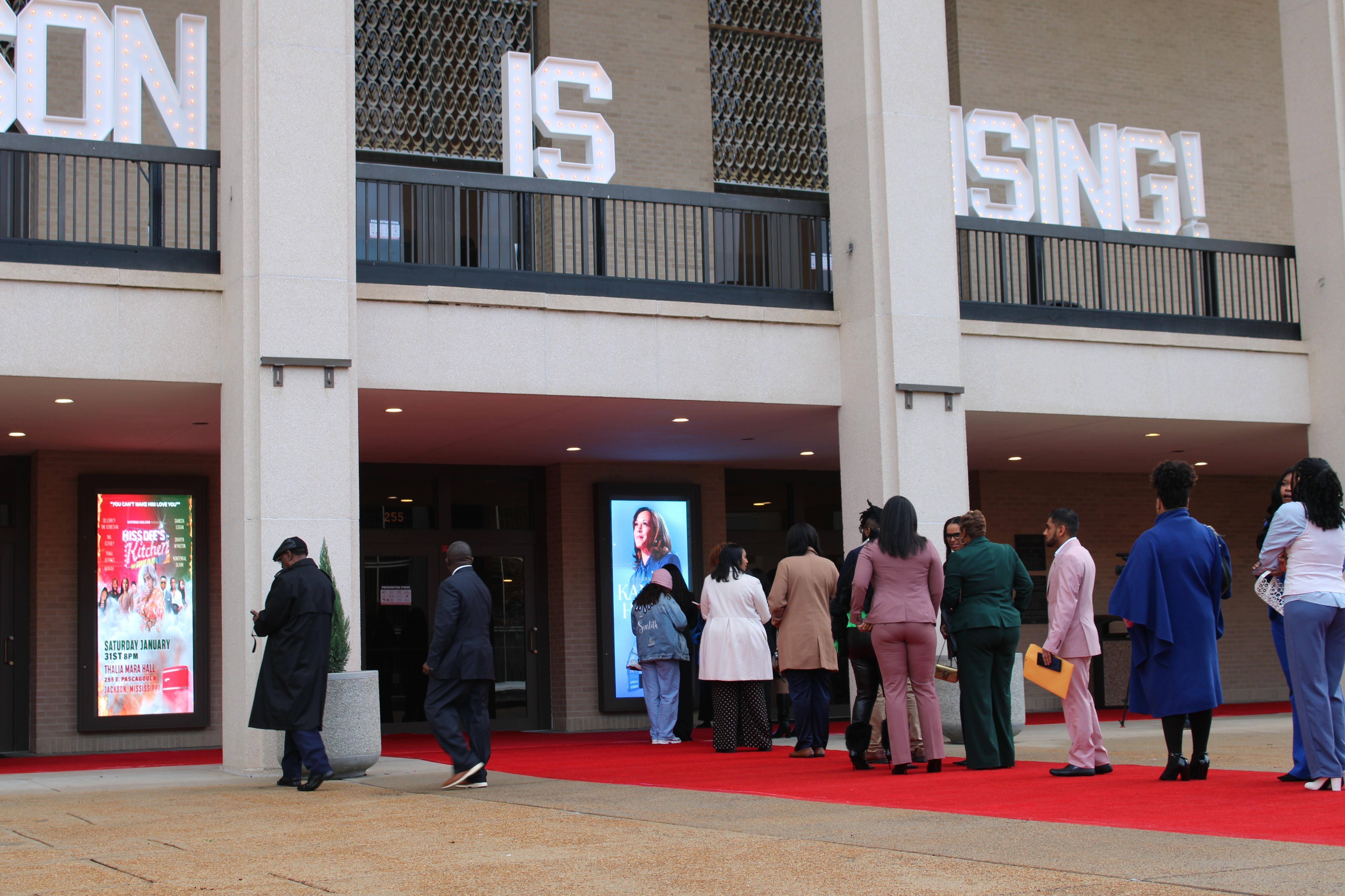 A VIP line for a "meet and greet" formed outside Thalia Mara Hall three hours before the main event. A line of people is seen on a red carpet in front of a concert venue with former Vice-President Kamala Harris's picture seen in the background.