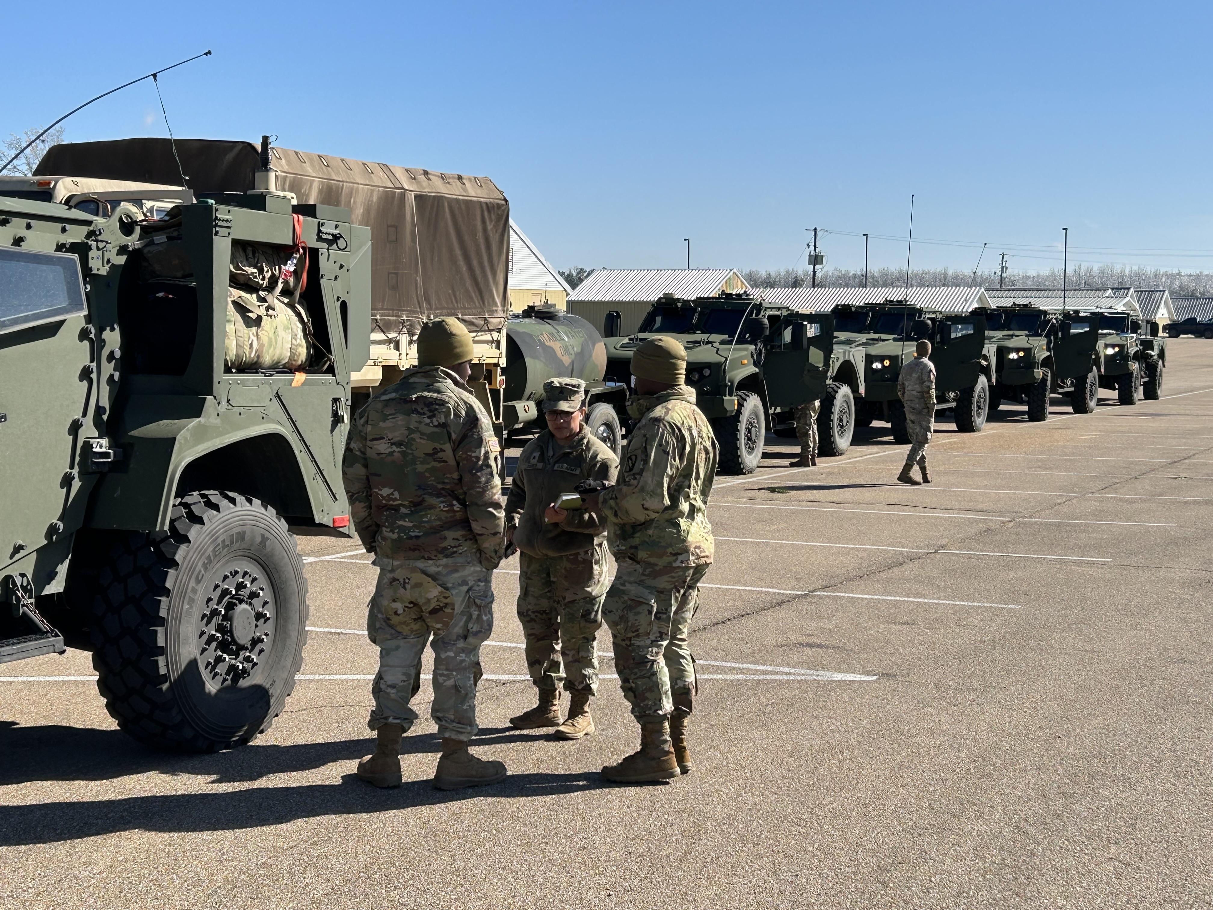 National Guard troops huddle in a group of three as military vehicles are seen next to them and in the background.