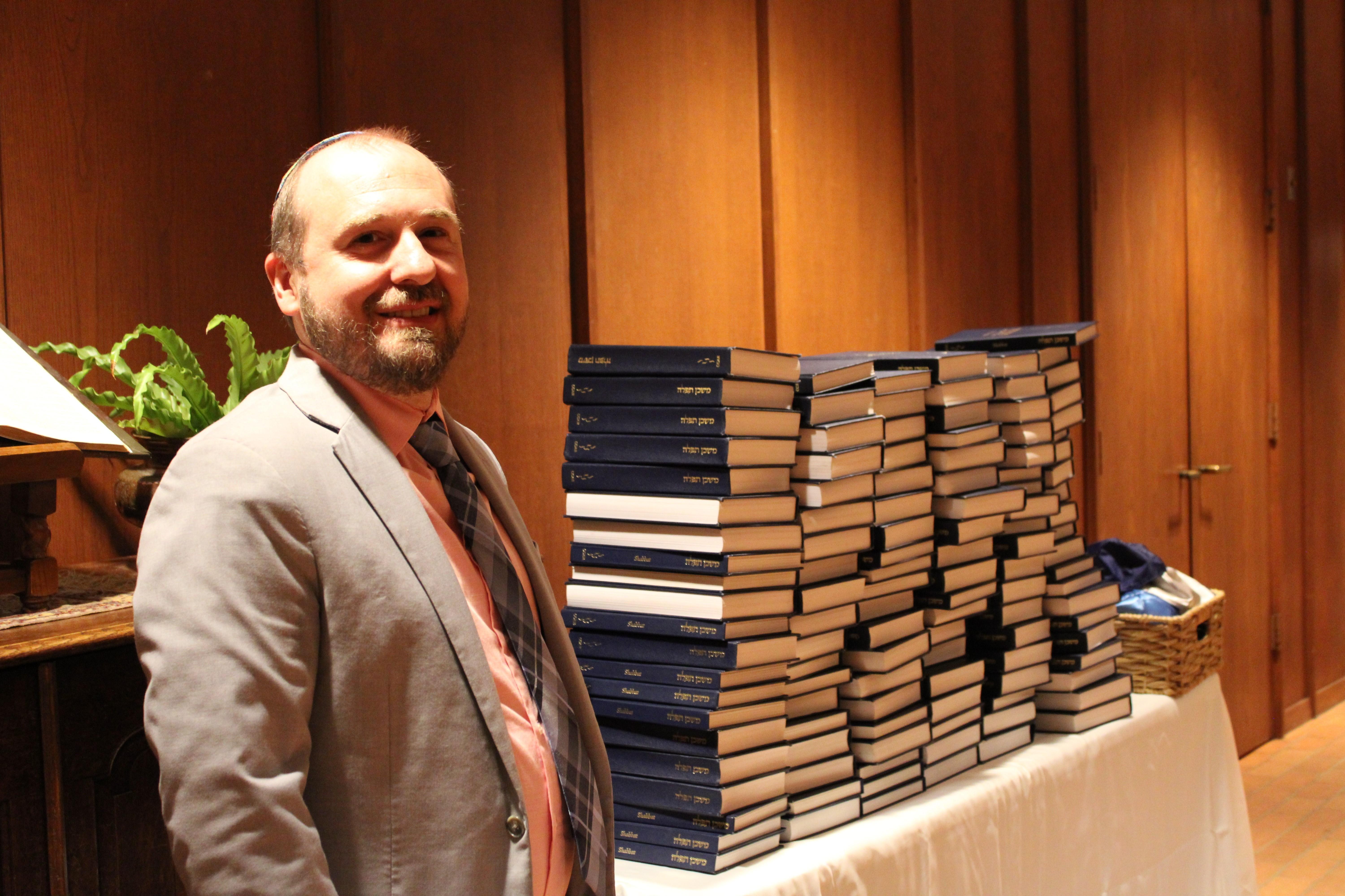 A rabbi smiles as he stands before a pile of blue Hebrew prayer books.