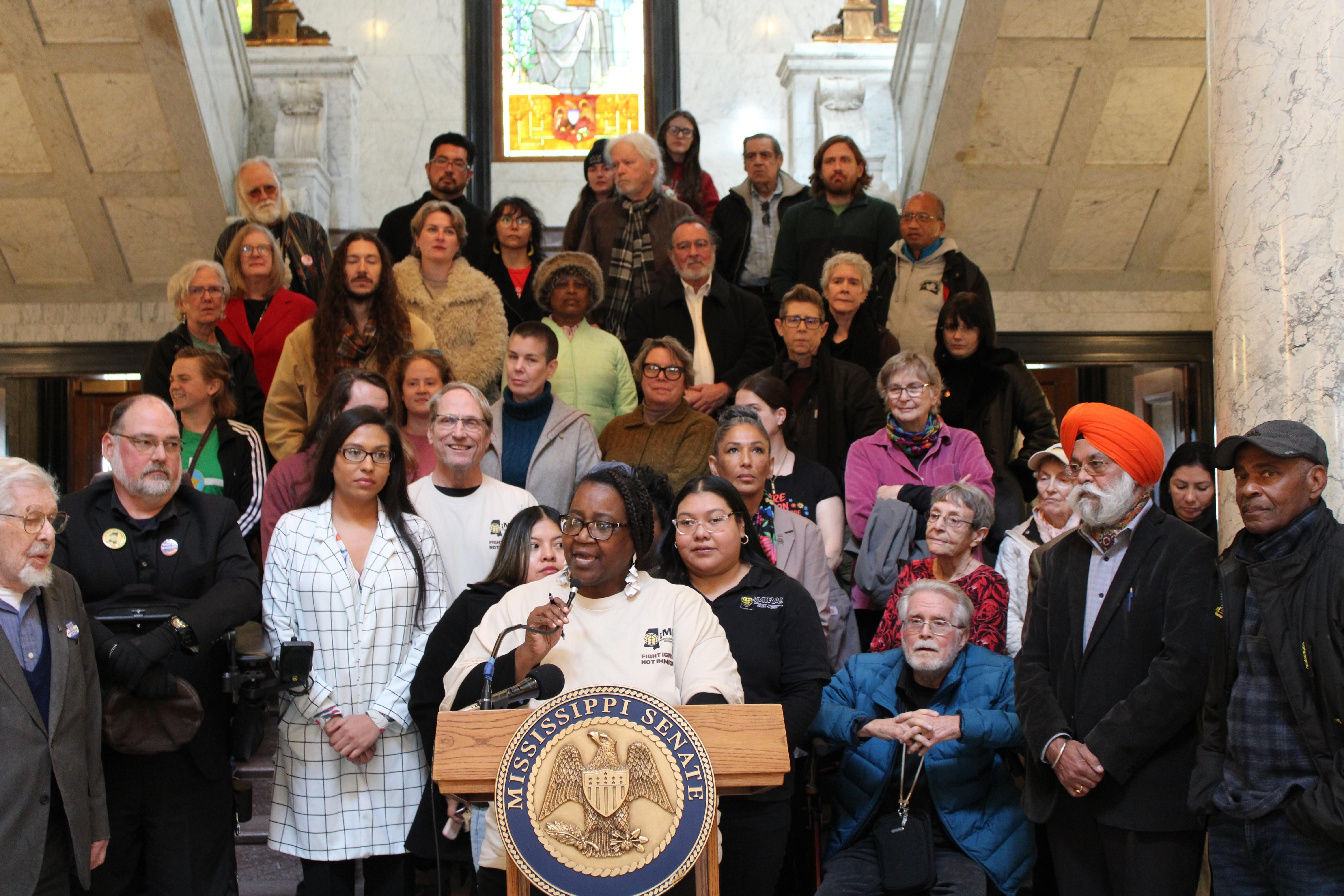 MIRA Executive Director Kathy Sykes speaks during a press conference at the state capitol building January 28, 2026 with more than two dozen people behind her.