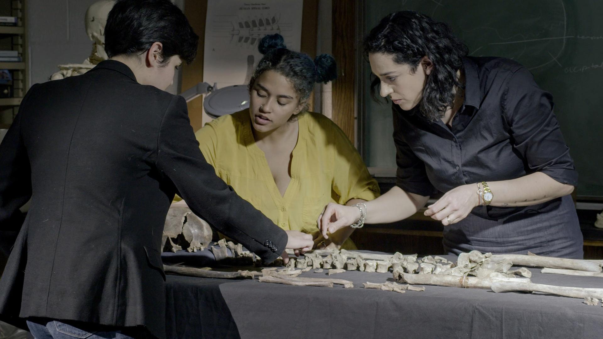 3 women study the bones of a skeleton that is laid on a table.