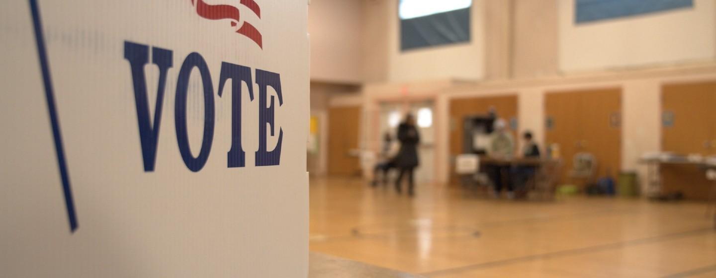 Vote sign in foreground with people handing out ballots in the background