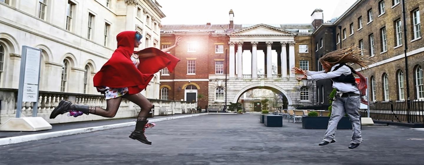 woman in red cape and man floating in air raising hands at each other in city center town square in front of old european buildings defying gravity