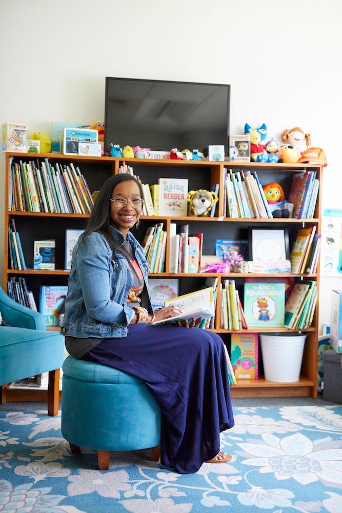 Mallory Mbalia sits on a blue ottoman with an open book in her office filled with books for children