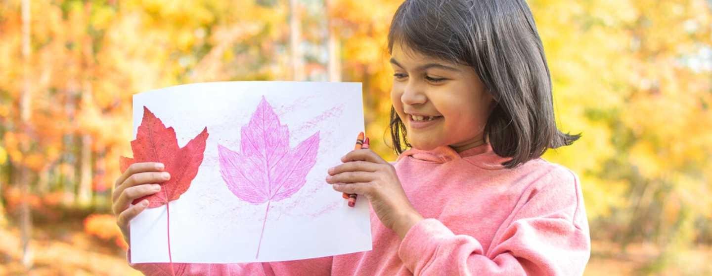 A young child about 8 years old holds up a color drawing of autumn leaves