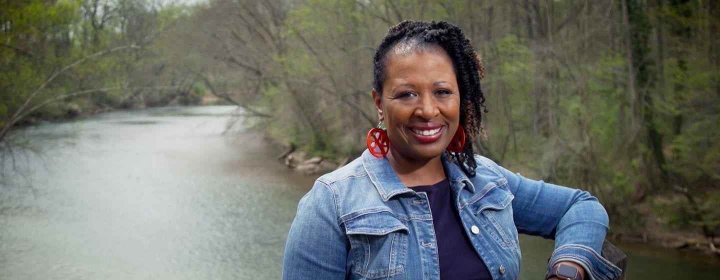 Deborah Holt Noel, host of NC Weekend in front of a river lined with freshly budding trees.