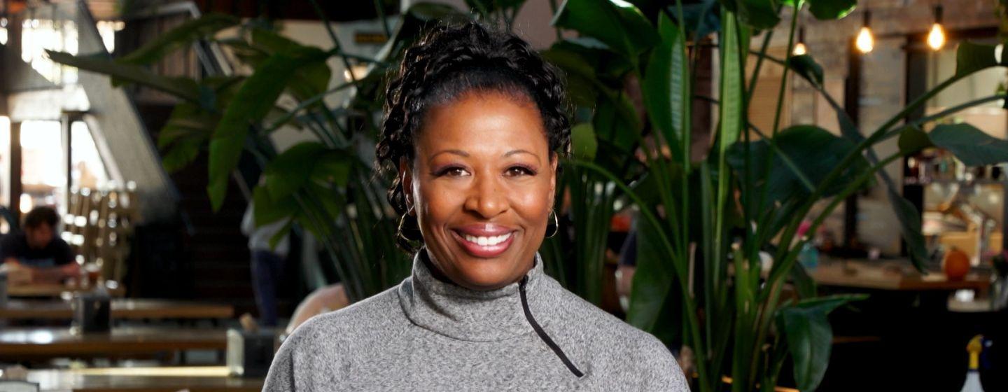 Host of NC Weekend, Deborah Holt Noel, wears a grey turtleneck in a restaurant full of plants.