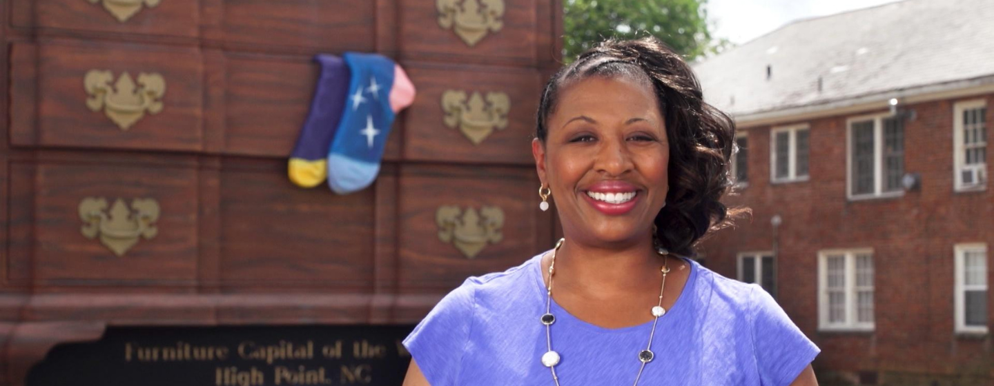 Host Deborah Holt Noel, a Black woman, stands smiling, wearing a purple shirt. She stands in front of a large chest of drawers with two blue socks hanging out a drawer.