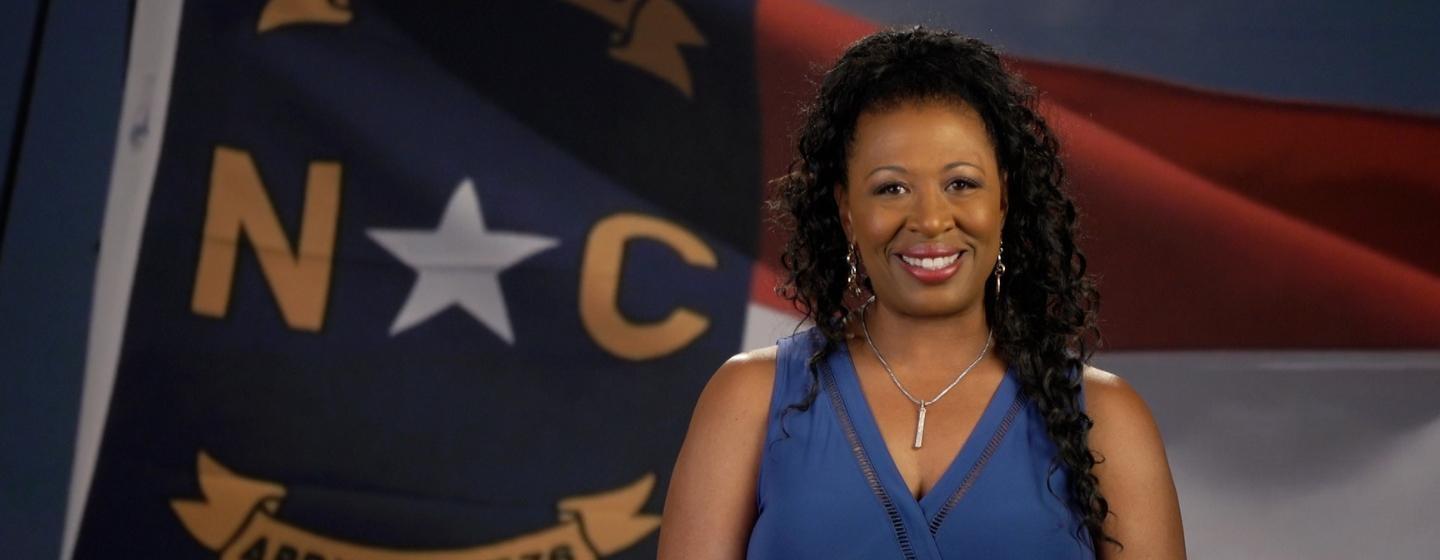 A photo of Deborah Holt Noel standing in front of the North Carolina State flag.