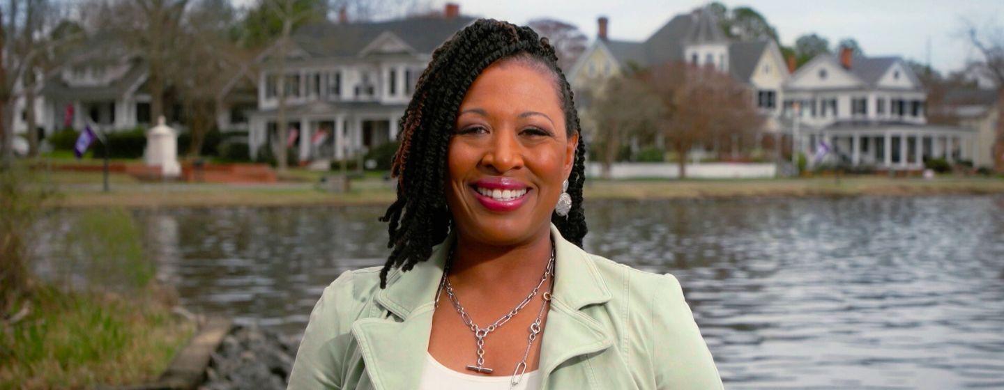 Deborah Holt Noel, host of NC Weekend in front of a lake with a a row of houses.