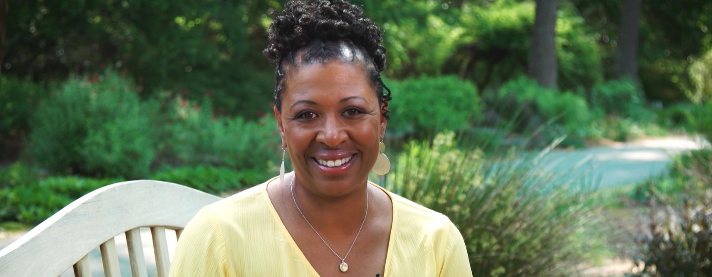 Host Deborah Holt Noel, a Black woman, sits smiling, wearing a yellow shirt. She is sat on a white bench in front of a small pond and various green plants.