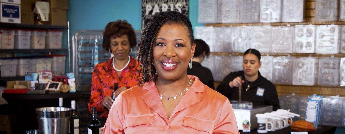 Deborah Holt Noel, host of NC Weekend in front of a chocolate shop counter with multiple people working behind it.