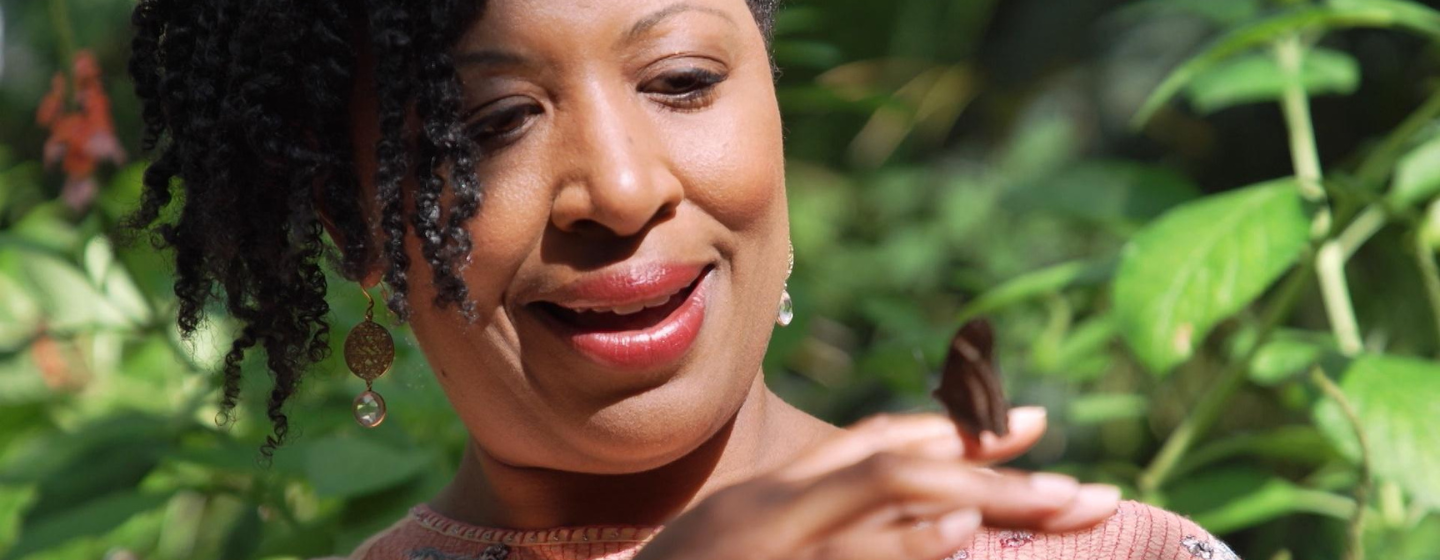 Host Deborah Holt Noel, a Black woman, is smiling, holding a butterfly on her finger. She stands in front of various green plants.