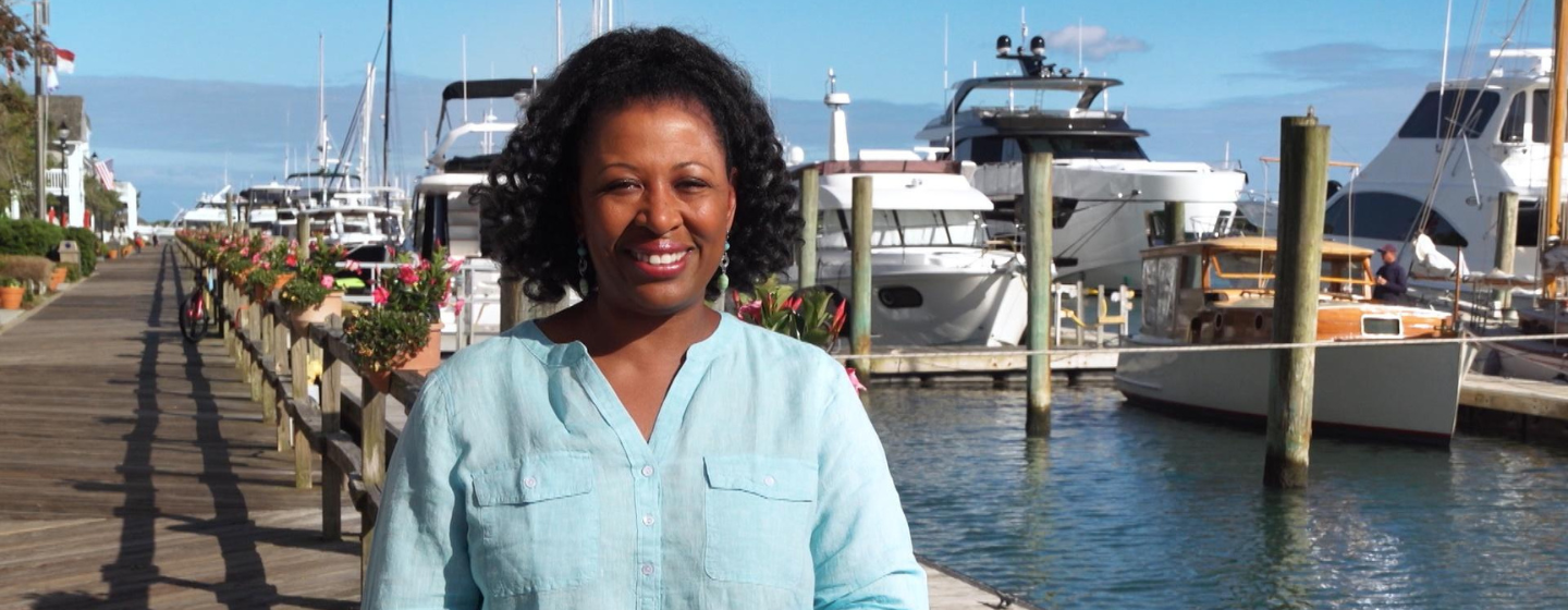 Host Deborah Holt Noel, a Black woman, stands smiling, wearing a bright blue shirt. She stands on a wooden boardwalk in front of a marina filled with white boats.