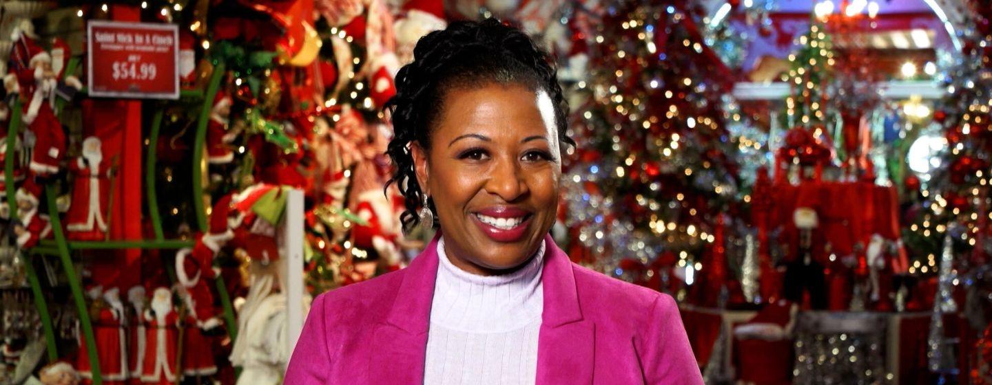 Host of NC Weekend, Deborah Holt Noel, wears a white turtleneck and bright pink blazer with a store full of winter holiday lights and decor behind her.