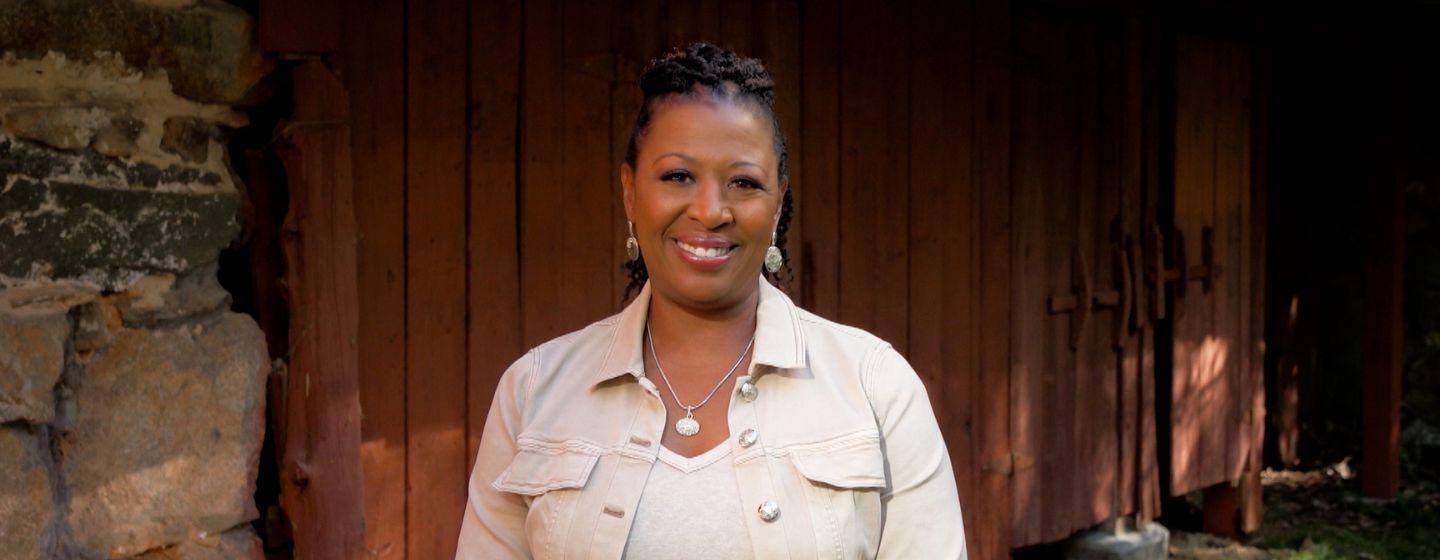 Deborah Holt Noel wearing a white v-neck tee with a cream colored jean jacket over it. She's standing in front of a red wooden door.