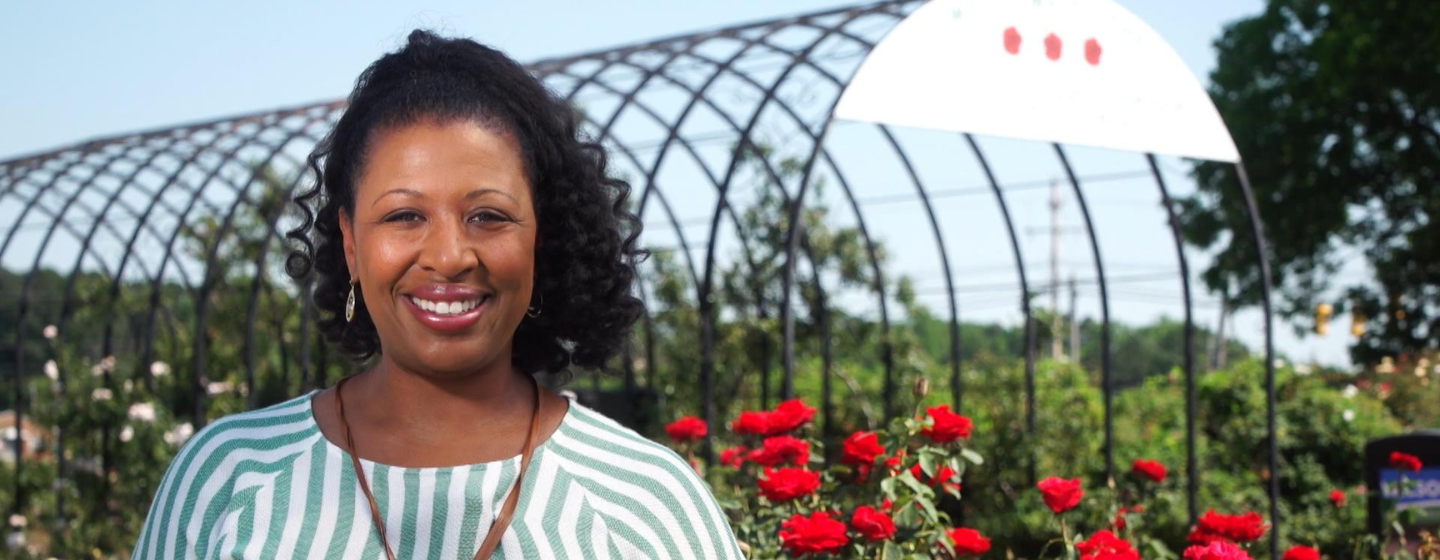 Host Deborah Holt Noel, a Black woman, is smiling, wearing a blue and white striped shirt. She stands in front of a large structure and red roses.