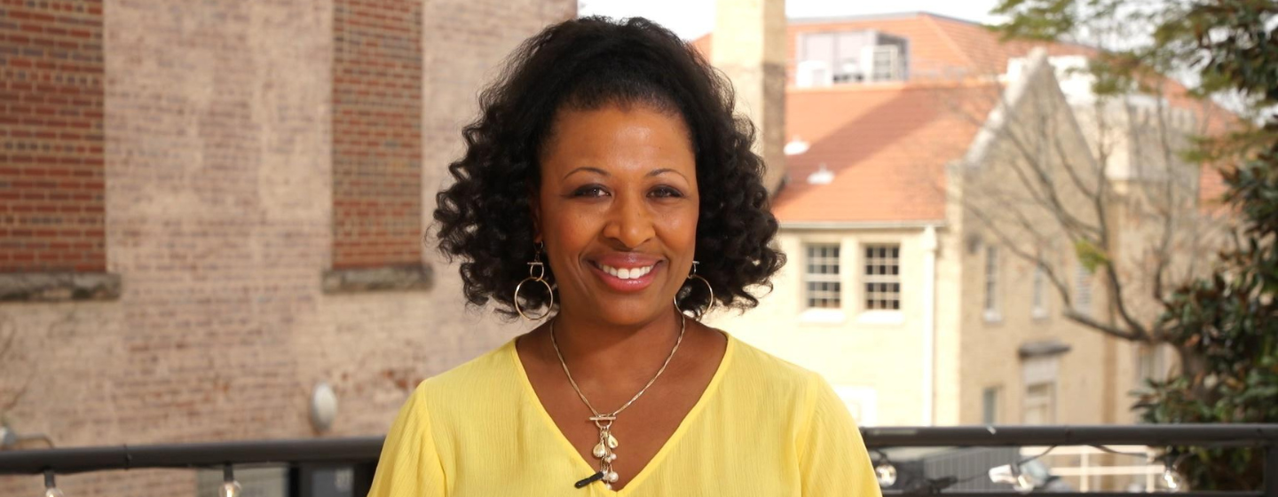 Host Deborah Holt Noel, a Black woman, stands smiling, wearing a bright yellow shirt. Behind her is a black metal railing and various brick buildings.