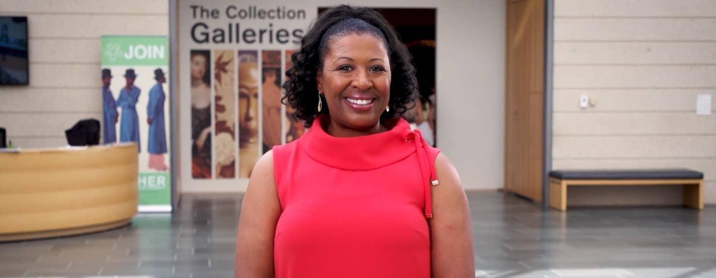 Host Deborah Holt Noel, a Black woman, stands smiling, wearing a bright red shirt. She is stood in front of an art gallery.