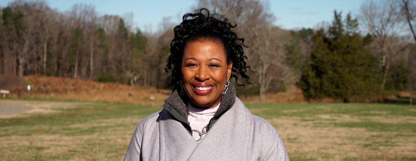 Host of North Carolina Weekend, Deborah Holt Noel wears a grey jacket and smiles while standing in front of a field.