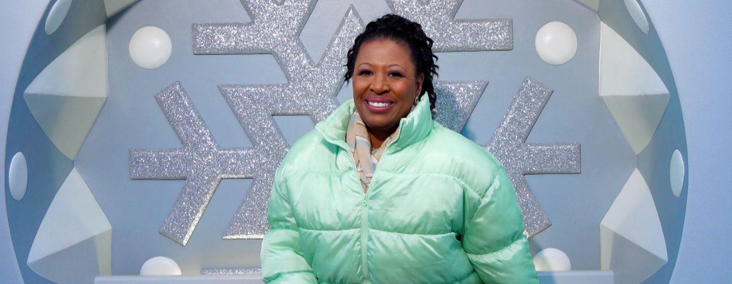 Host of NC Weekend, Deborah Holt Noel wears a pastel green puffer jacket in front of a snowflake sculpture.