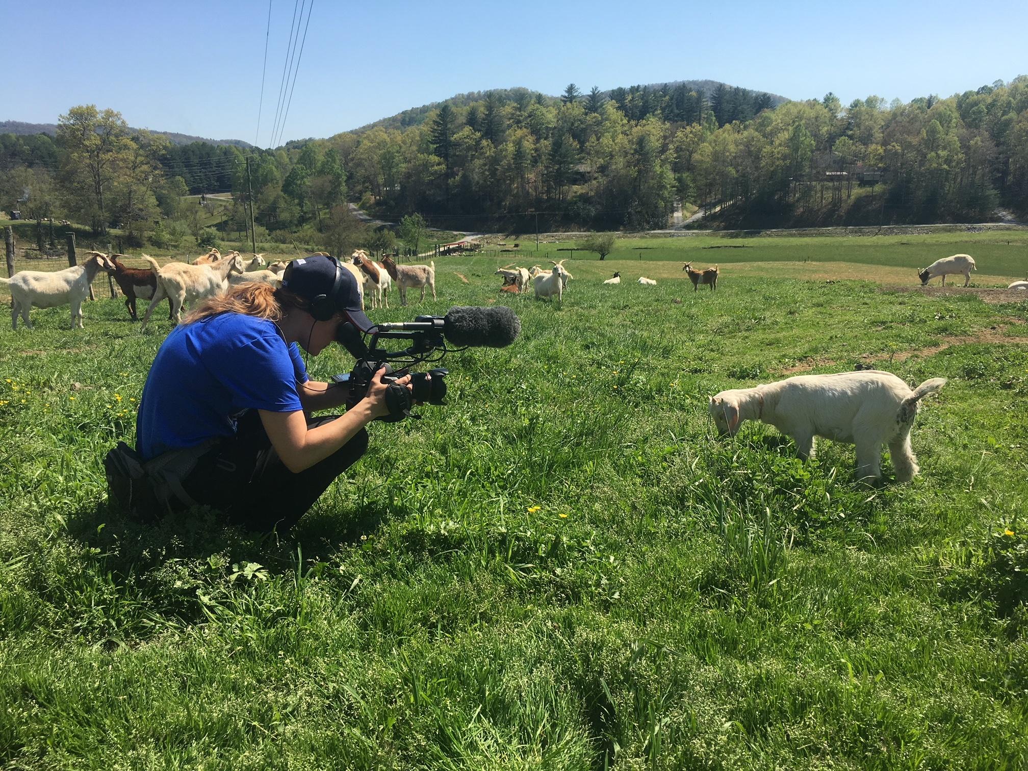 Blonde crew member sits in front of goat while holding up a camera