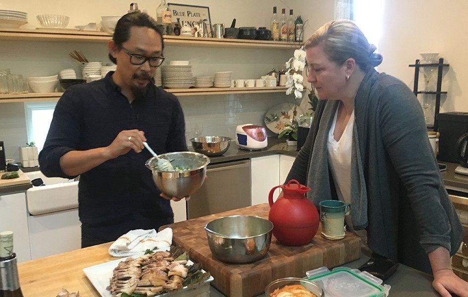 Chef Ashley Christensen watches Joe Kwon prepare food in a kitchen. She leans on a counter filled with cooking supplies and food.