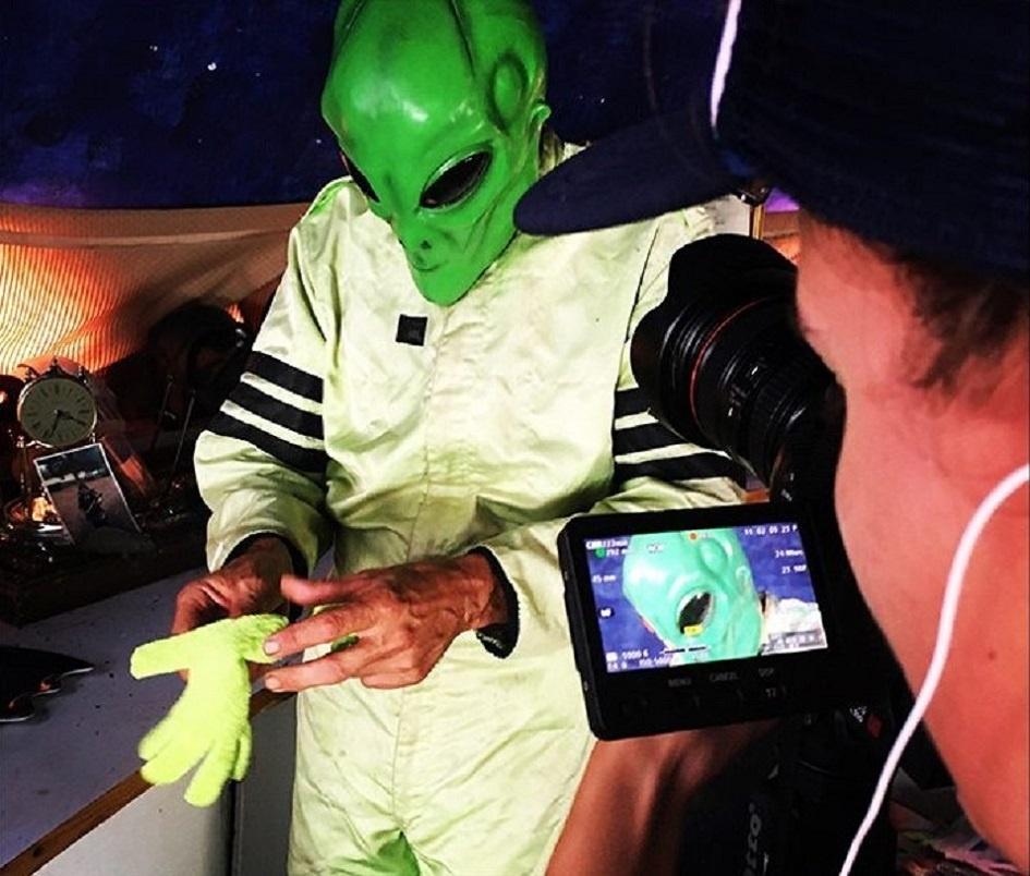 A man puts on a green costume, green alien mask, and green gloves. A cameraman films a close up.