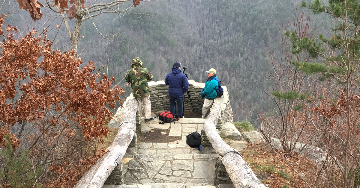 Three people standing near the edge of a brick-layer ledge. Passed that is a mountain range full of trees