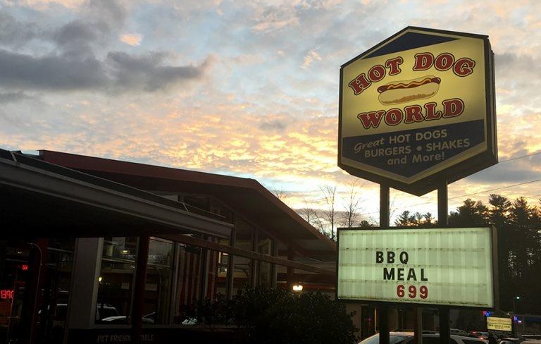 Image of the outside of hot dog world featuring the street sign on display and lit up. Sign says hot dog world with picture of a hot dog in the middle. A board is placed under it advertising a BBQ meal for six dollars and 99 cents