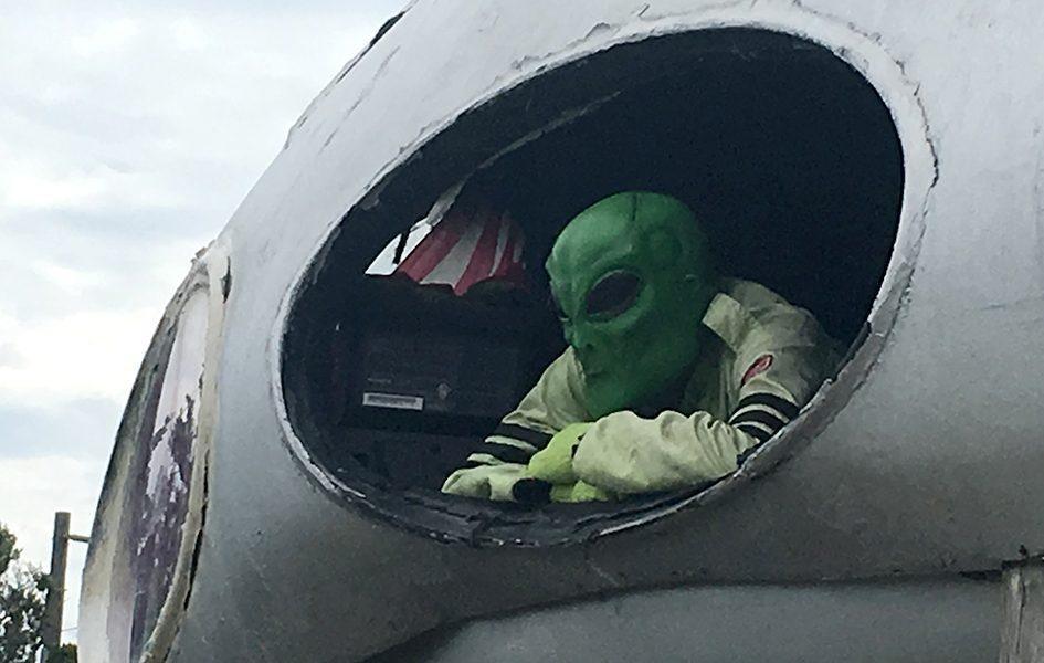 A man in a green suit and green alien mask rests his head on his folded arms. He peers out of a round window.