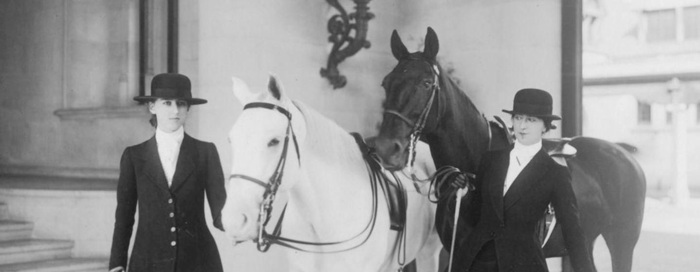 A black and white image of two women dress in dark clothing stood at the entrance of a building with two horses, one dark and one white.