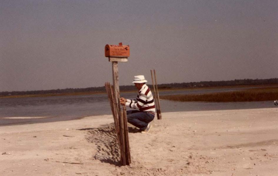 person fixing a post in the sand with a mailbox on top and a body of water in the background