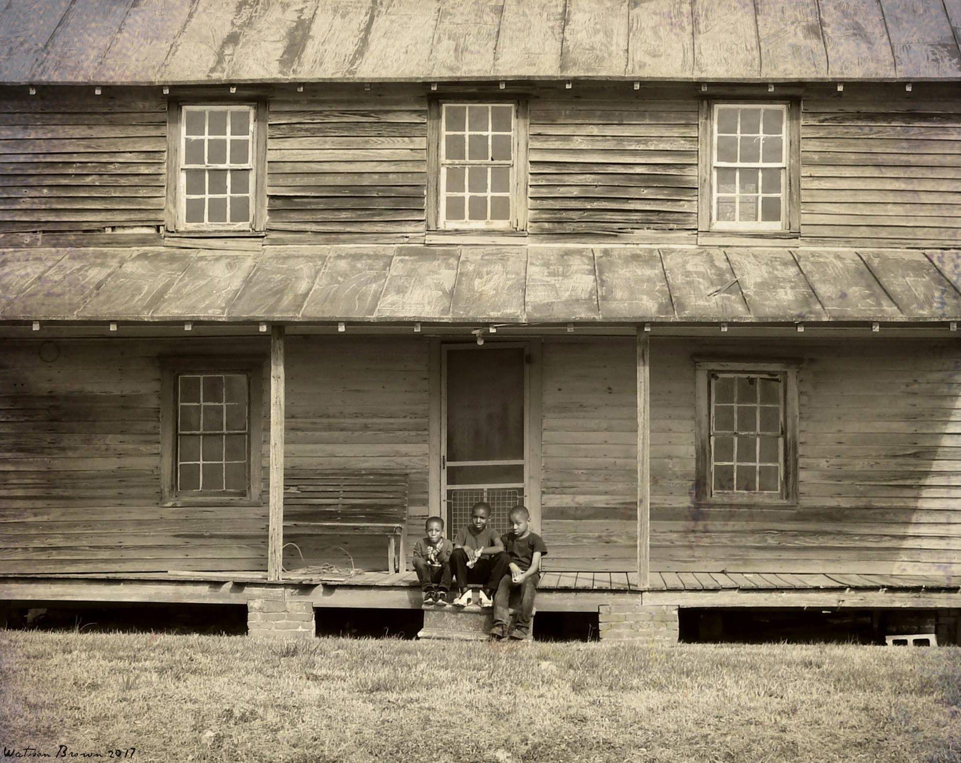 Three young Black boys sit on the edge of the porch of a historic house